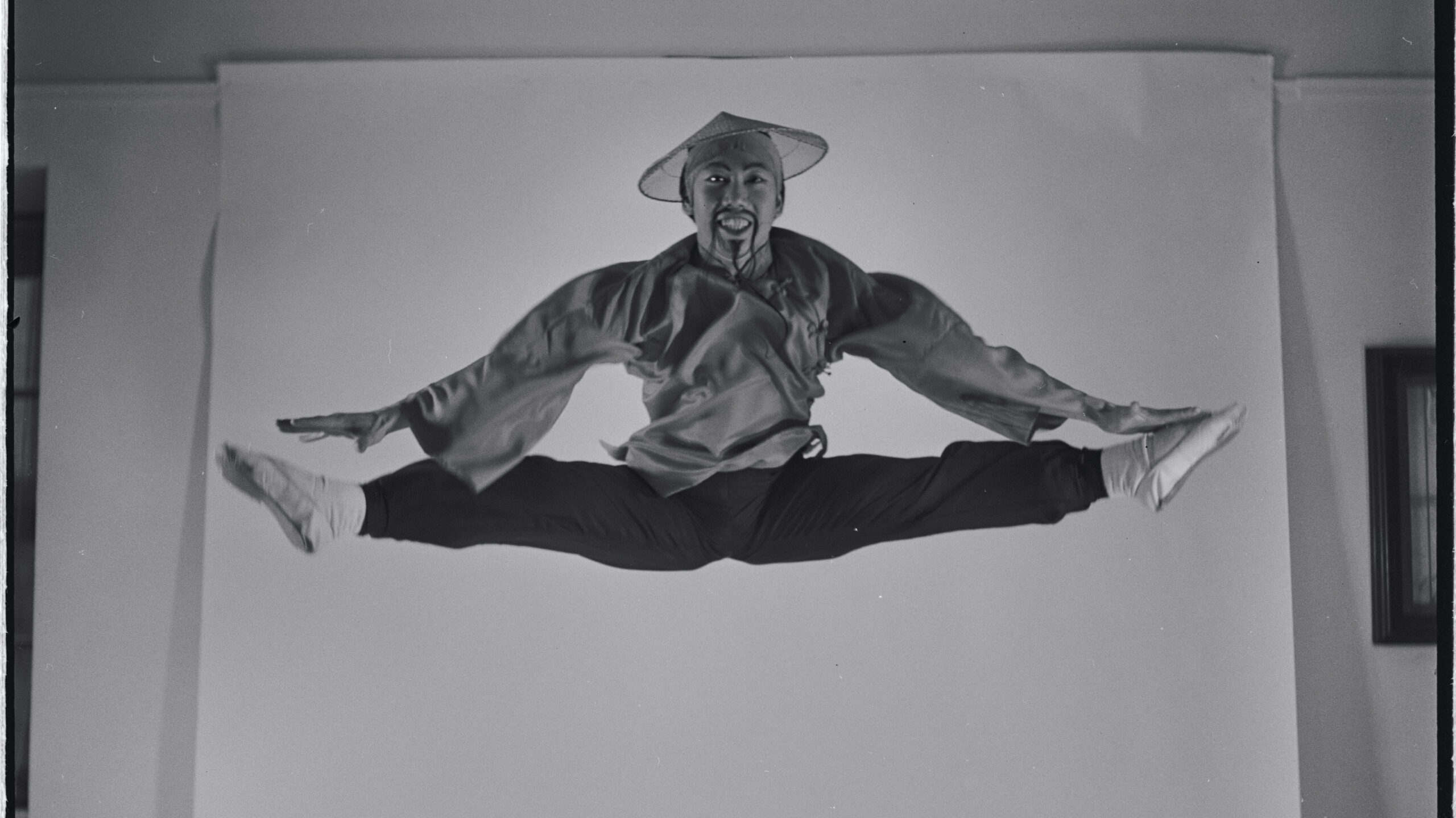 Black-and-white photo from 1954 of George Lee, a Chinese man, performing a mid-air jump split. He wears theatrical costume and a painted beard, with exaggerated facial features in yellowface makeup.