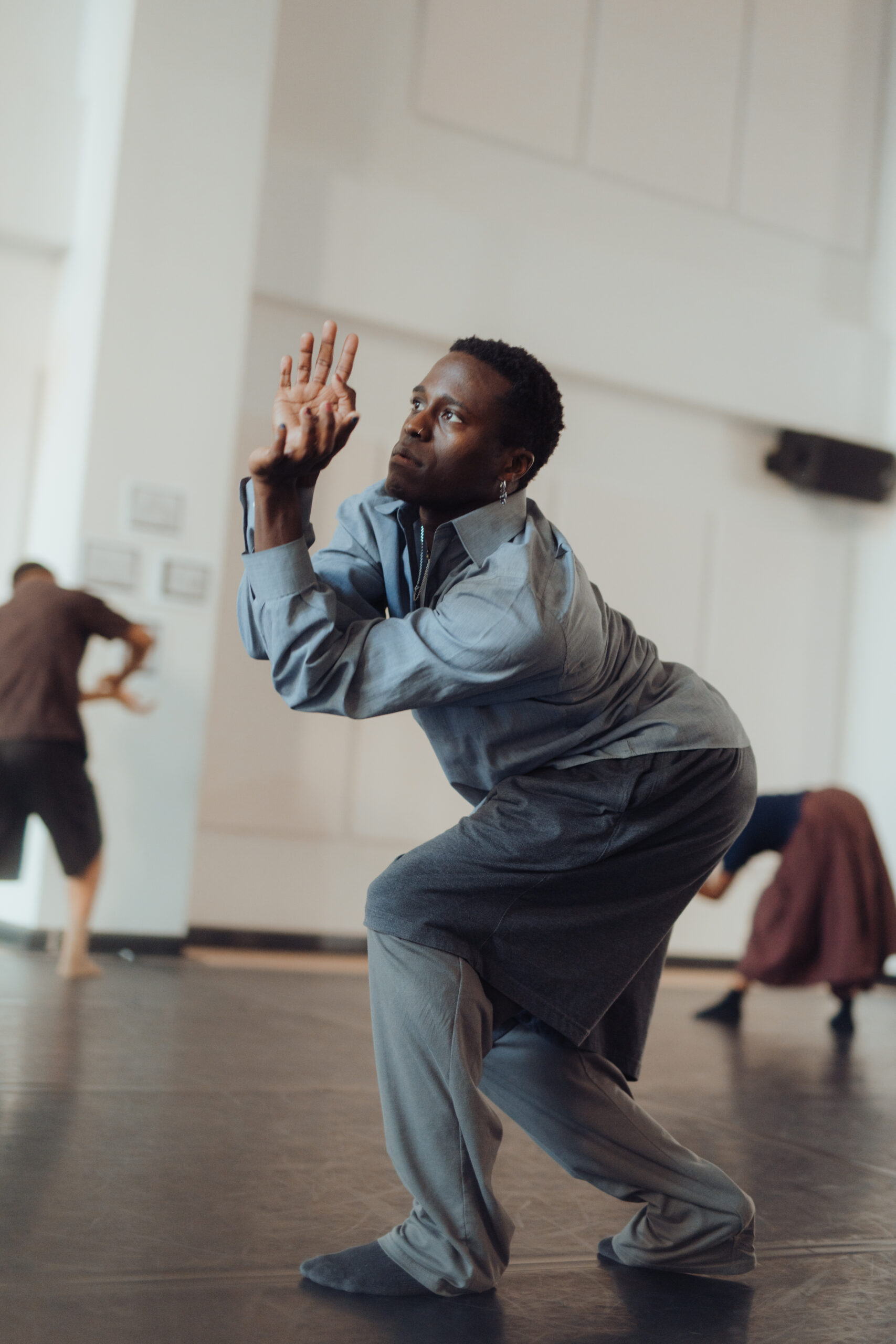 A dancer wears layered gray clothing as they bend forward, somewhere between a crouch and a lunge. They push their palms out in front of their face, with fingers gently curved, as if pushing something upward into the air.