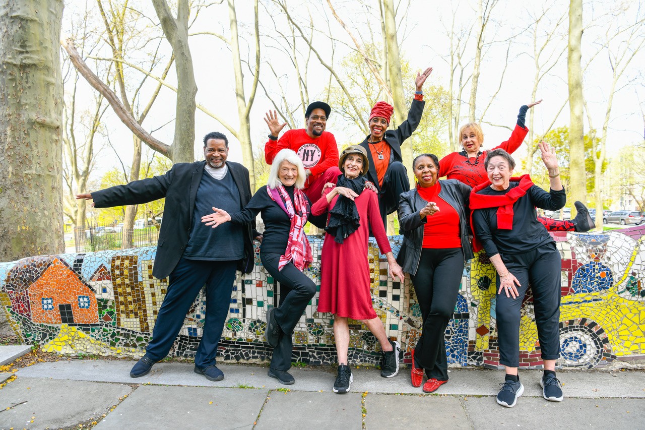 Eight senior dancers pose in front of a mosaic wall and a park. Their smiles wide, they open their arms, wave, and one points towards the camera. They are wearing black and red.
