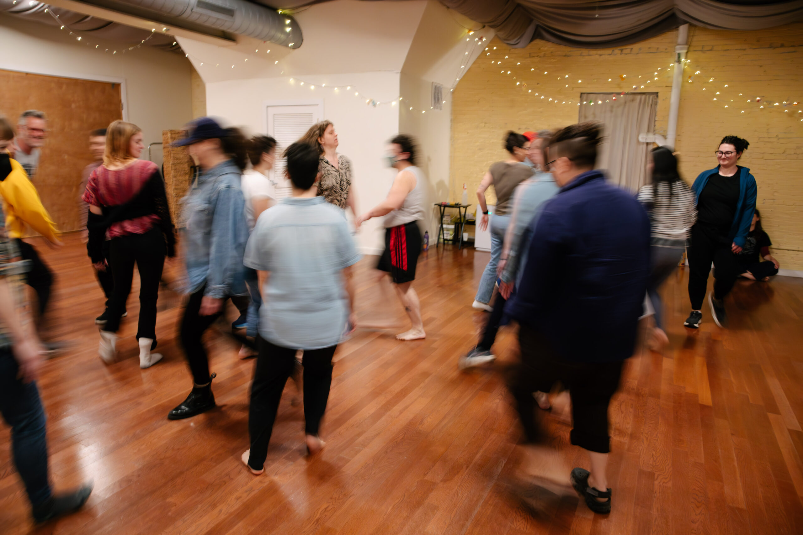A crowd of audience participants walks through a warm room with a wood floor and string lights in the background. Mid-motion, the camera blurs most of the participants as they smile and walk in-between and around each other.