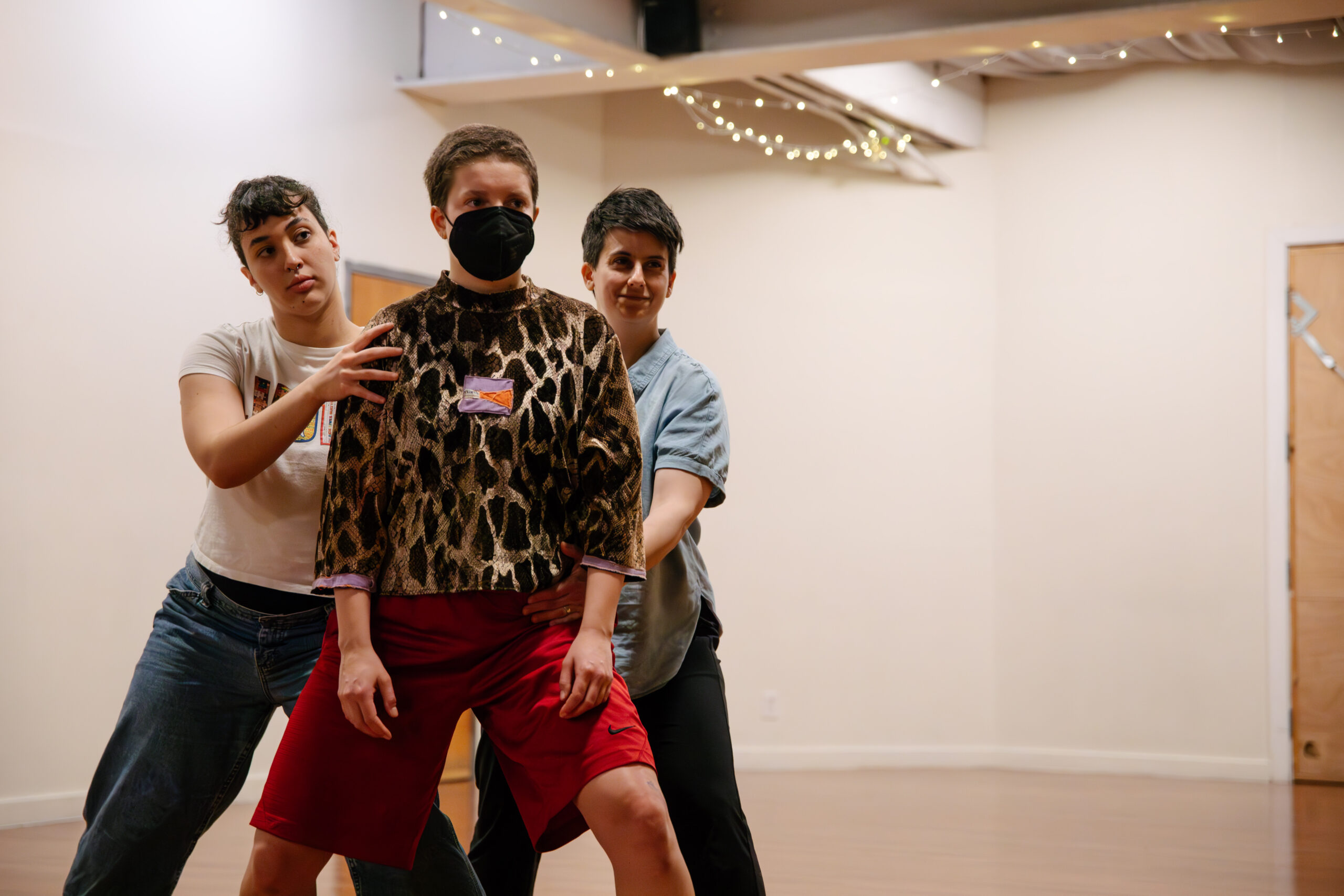 Three performers stand in a triangle in Studio 34. The camera blurs the background and focuses on their upper torsos and faces. The two dancers in backwear jeans and t-shirts; one laughs and the other holds a serious expression, bracing for impact. Together, they support the front dancer’s hips and shoulders. This third performer looks expectantly forward for the shove of another performer not in the photo.