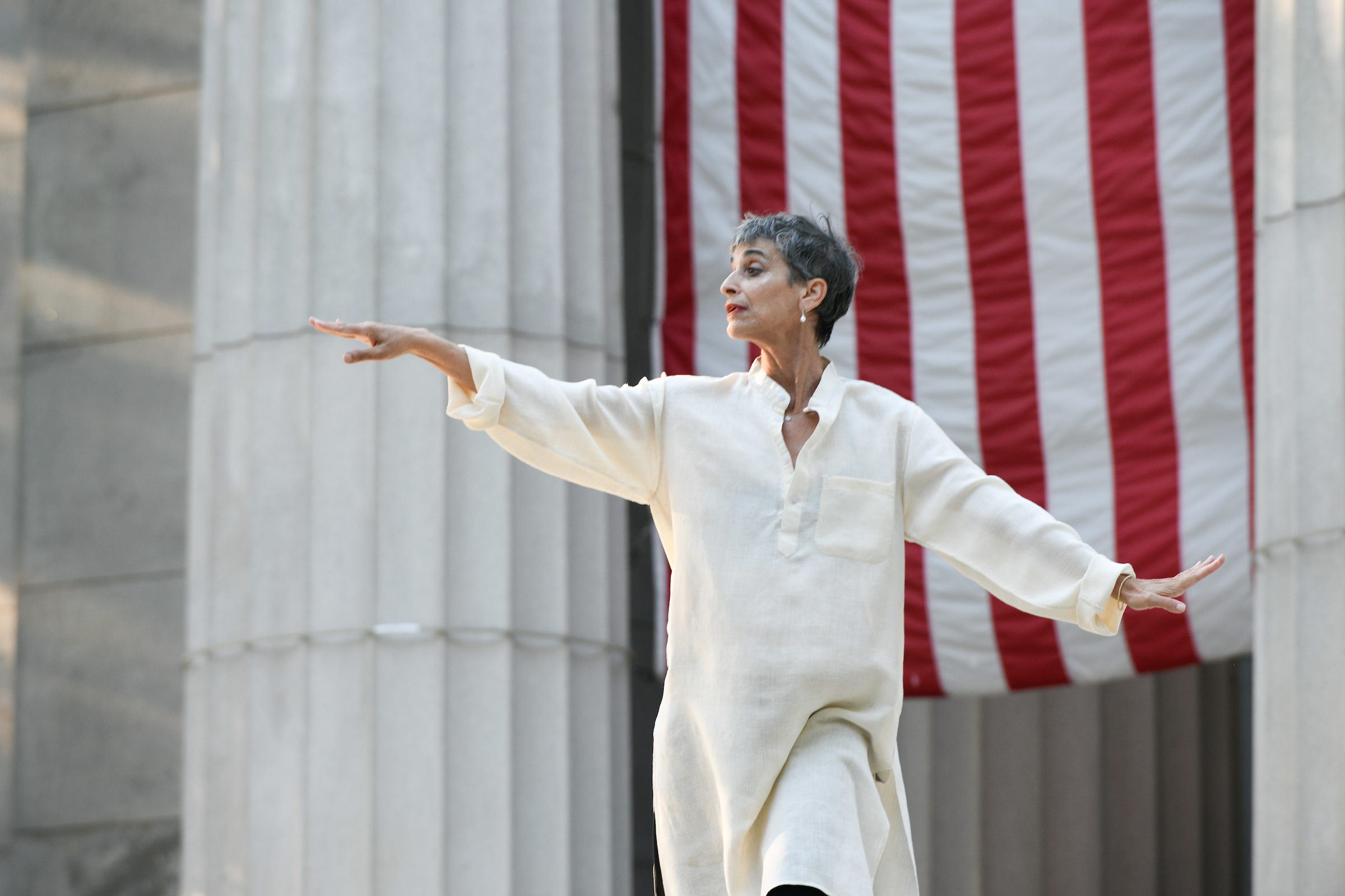 A woman with short cropped hair wearing a long white tunic balances on her left leg in front of a large stone pillar and a section of the American flag. She looks over her extended right arm, her wrist gently lifted.