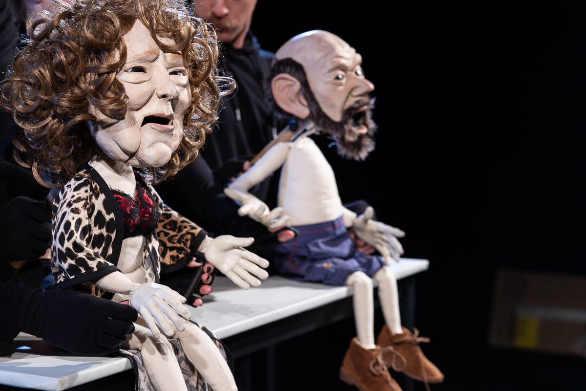 Two human-shaped puppets seated side by side on a platform, their legs hanging over the edge and their arms extended forward as if gesturing outward. Both puppets feature detailed facial expressions with open mouths. The puppet on the left has curly hair and wears a cheetah-print robe layered over a red bra with black lace, while the puppet on the right appears bald with facial hair and is dressed in shorts and shoes. Puppeteers dressed in dark clothing are partially visible behind and around the figures, with their hands engaged in supporting and controlling the puppets. A dark background and focused lighting emphasize the puppets’ faces, clothing, and posture, creating strong contrast and directing attention to the figures.