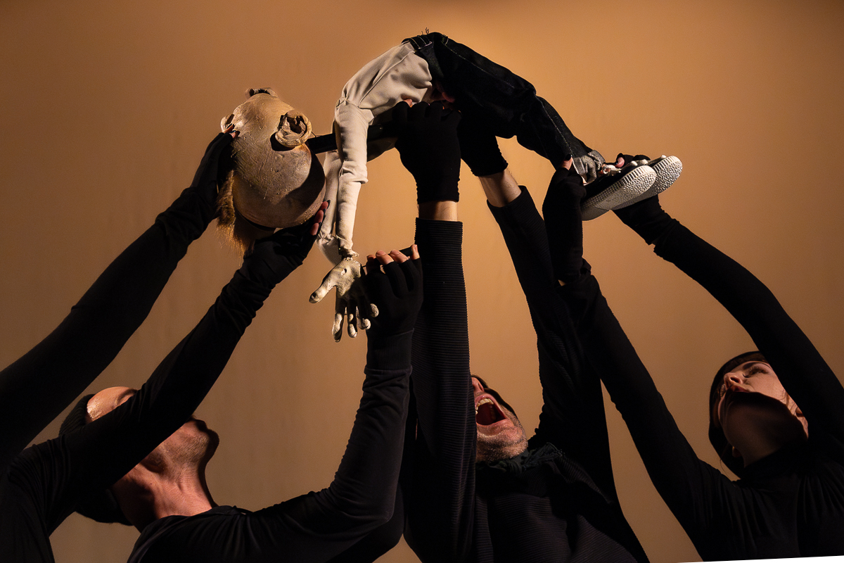 Four puppeteers dressed in black, standing close together with their arms raised as they support a small, human-shaped puppet above them. The puppet is shirtless and wears denim jeans and sneakers. It is held horizontally in midair, with its arms extended outward and its chest and abdomen lifted highest, creating a sense of suspension. The puppeteers are only partially visible, their faces and arms angled upward toward the puppet. A plain, warm-toned background and controlled lighting create strong contrast between the figures and the setting.