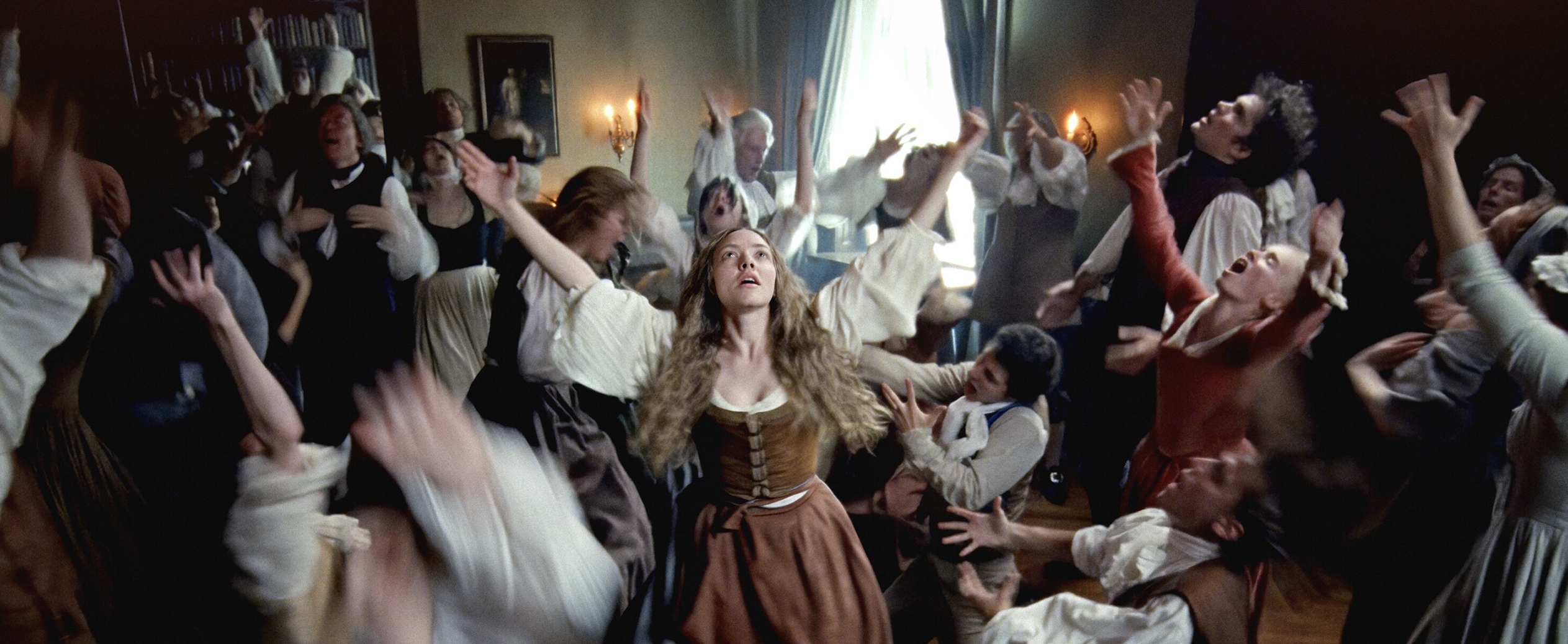 A scene from the 2025 film, The Testament of Ann Lee: Deep in the throes of ecstatic worship, Ann Lee (Amanda Seyfried), leads her congregation in an endurance challenge of dance, song and prayer. Men in powdered wigs and billowing sleeves and women in long dresses toss their heads back, open their mouths, and raise their arms. Two men near Lee kneel and stretch their arms out toward her. Lee is the only character fully in focus. Her blonde hair flies in front of her shoulders, chest heaving, as she raises her arms and eyes toward the Divine.