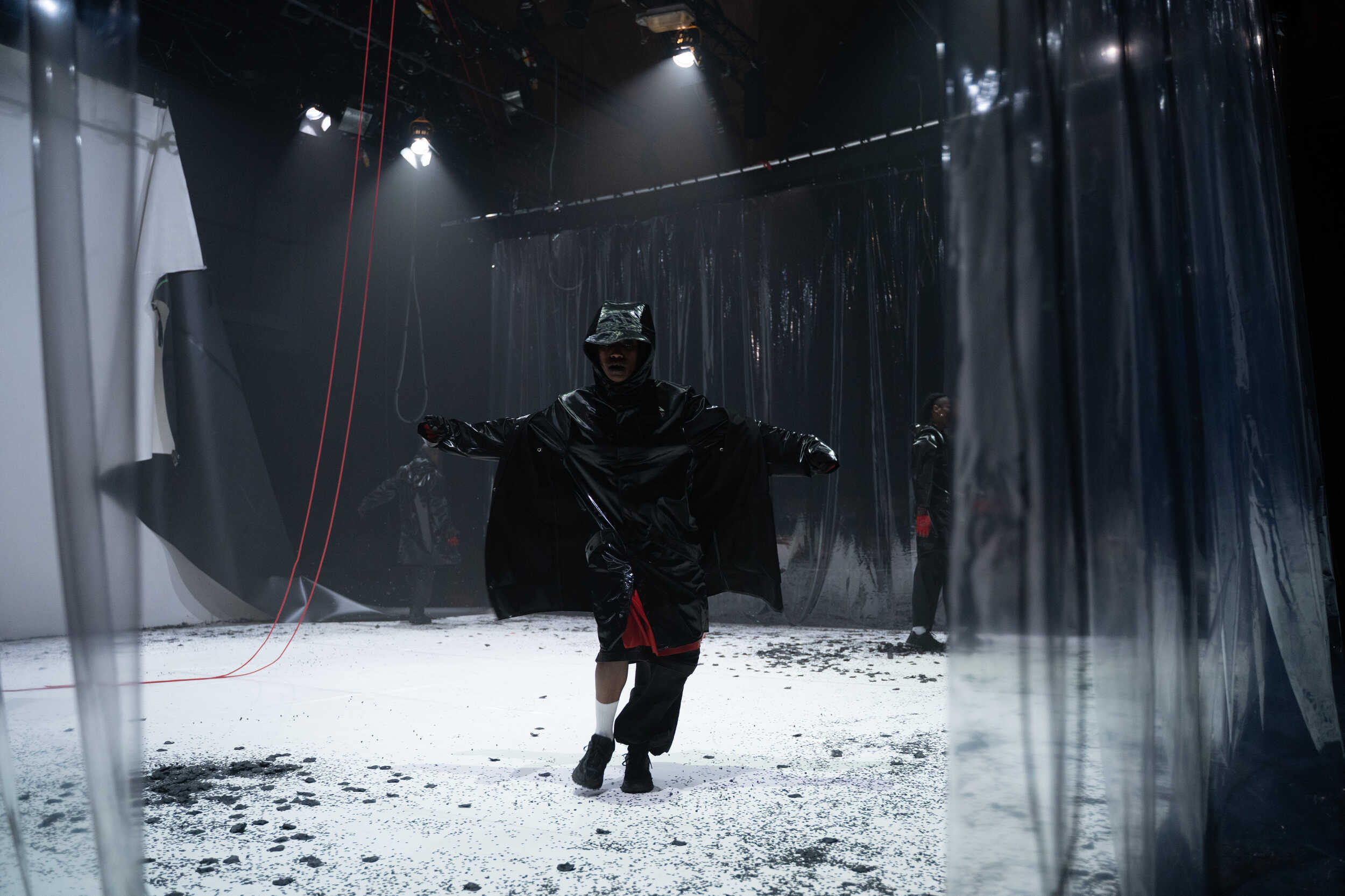 Mulunesh, a Black woman in a thick, hooded raincoat, stands crookedly with her weight shifted over one foot. Her arms are lifted out from her sides and her hands are in fists. She is lit with harsh, bright lights, and boxed in on three sides with heavy transparent plastic. Behind her, a sheet of white marley and two red cables dangle limply, as if caught mid collapse. The floor beneath her feet, made of the same white marley, is spotted with piles of black paper confetti.