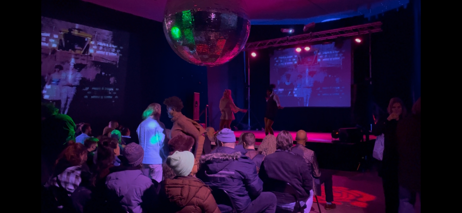 The audience is awash in purple; the room is dim and the discoball floats overhead, reflecting green and red and purple. There is projection on the back wall of the stage that is purple with blurry outlines of people, and another projection on a sidewall with people dressed in white in a dress and clownish costume. The audience mills about, some with drinks in hand. A performer in a black dress sings into a microphone onstage while someone else appears to be facing them, speaking to them.