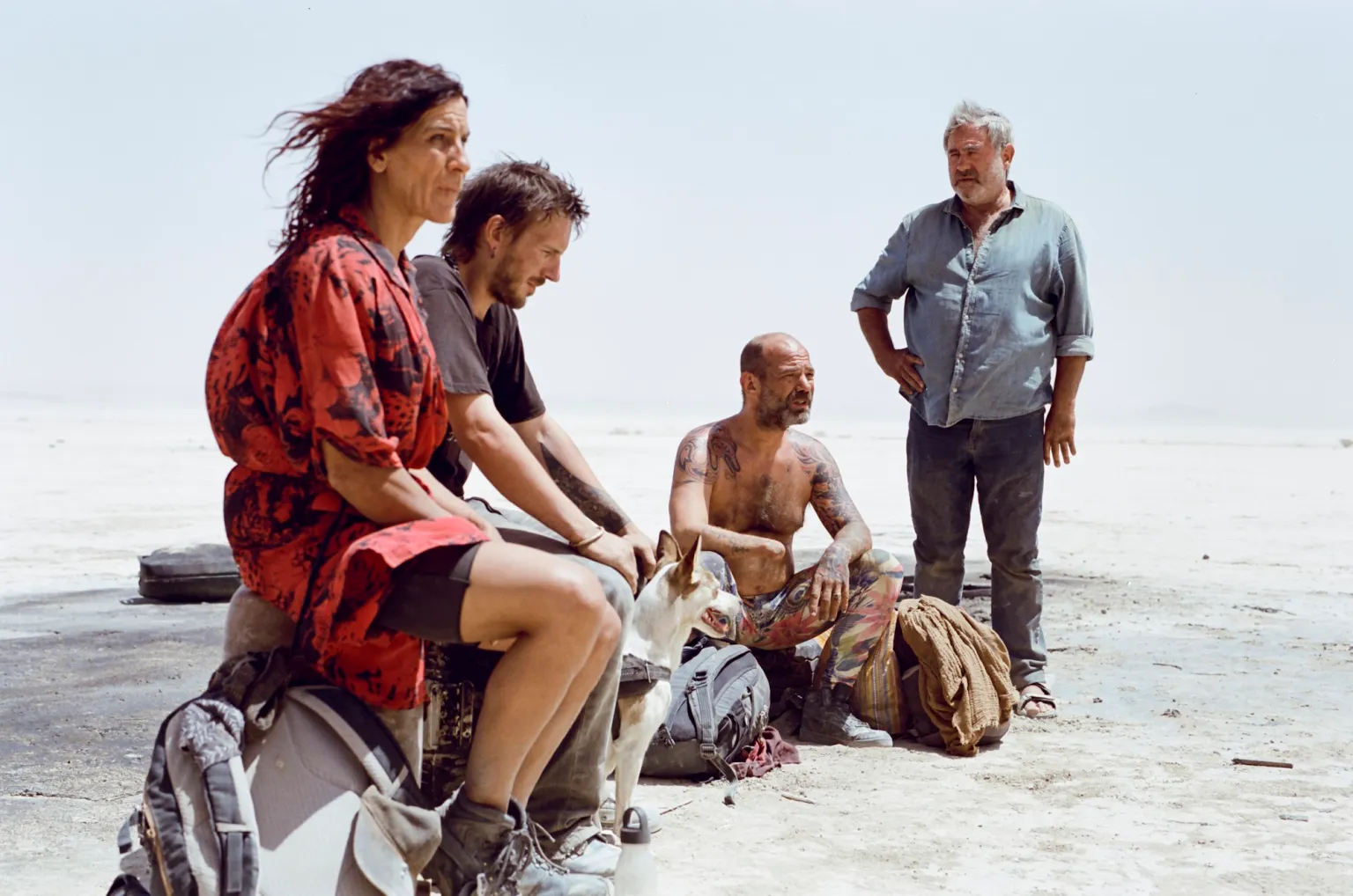 Four people in a desert. On the left, a woman with black hair in a red dress sits, a dusty backpack at her feet. Next to her, a bearded man with short brown hair sits looking down, his hands on his knees. To his left is a second man, bald and bare from the waist up, with tattoos covering his left arm and his right arm missing his hand. To his left is a third man, standing, with one hand on his right hip. He is older than the rest, has a white beard, and wears a light blue shirt.