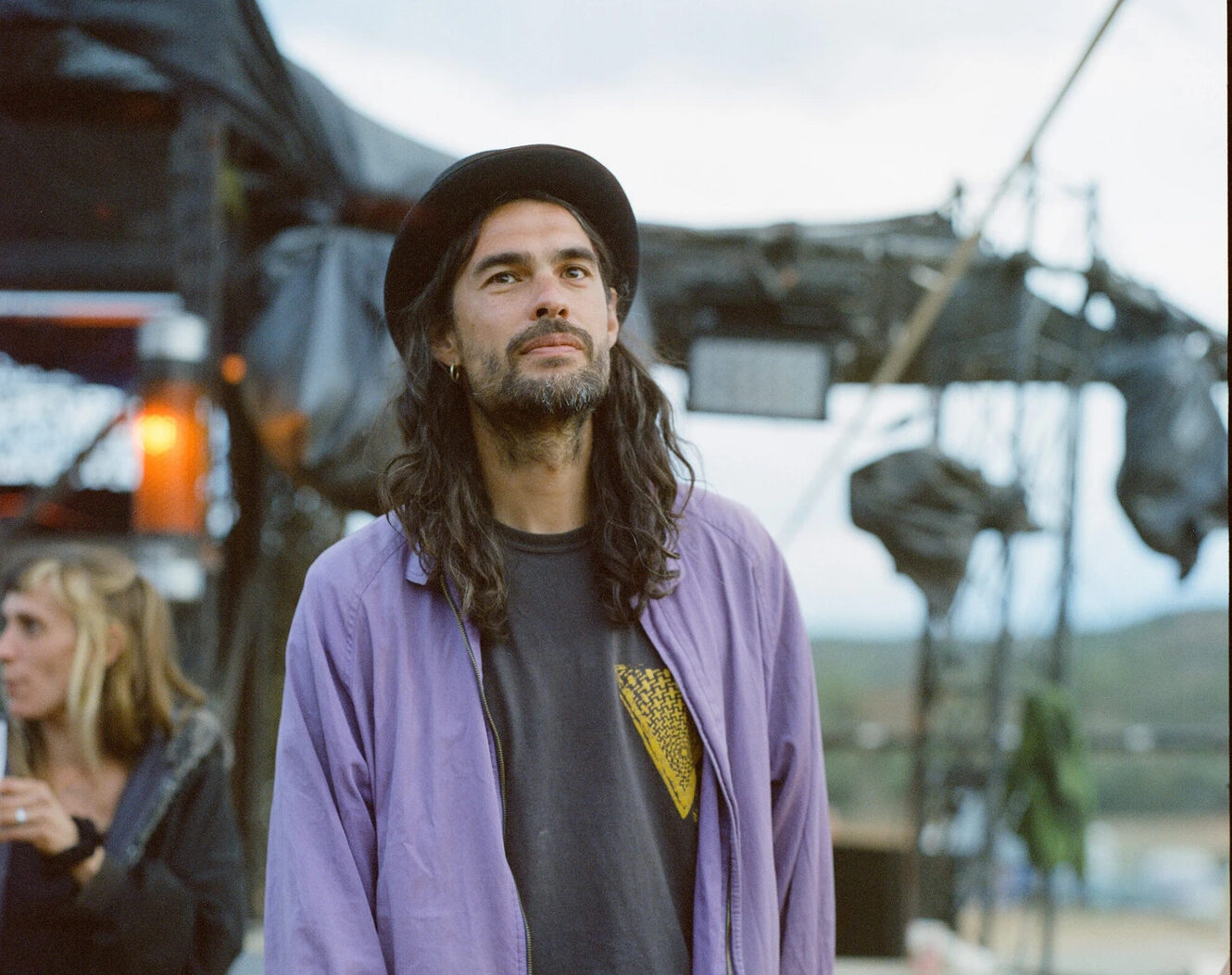A young man stands relaxed in front of what looks like some kind of set in the middle of the desert, a woman (out of focus) with blonde hair stands behind him to his right. The man has long black hair that trails down to his collarbones, and matches the color of his beard. His hat is tilted towards the back of his head, and he wears a light blue jacket over a black tee-shirt, and leopard-print pants.