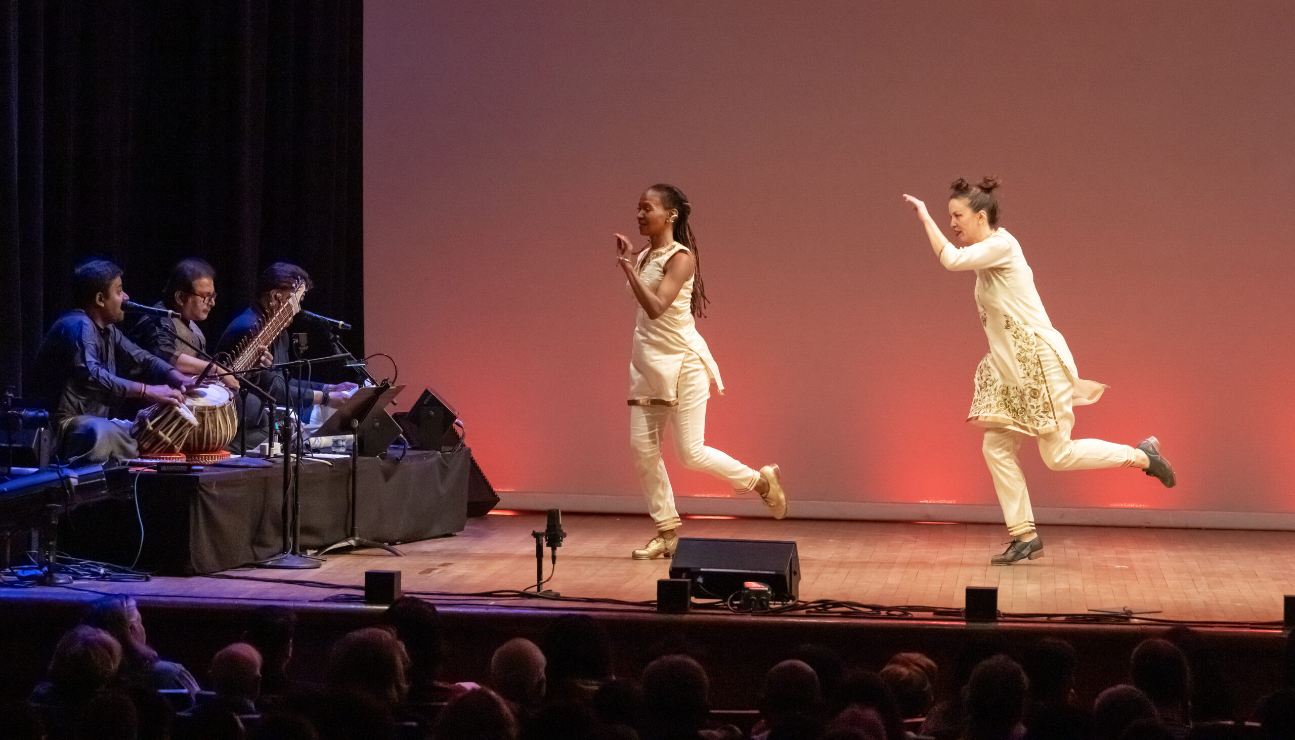 From left to right, dancers Dormeshia, Rachna Nivas, Rukhmani Mehta and Michelle Dorrance. They are in motion. Dormeshia and Dorrance wear white pants, thigh length white tunics, and tap shoes. Nivas and Mehta wear white leggings, long white dresses with golden details on the skirts and bodices. They have bands of bells around their ankles and are barefoot. The tap dancers have a quality of bending and sending energy into the floor. The Kathak dancers are lifted, arms raised, poised.