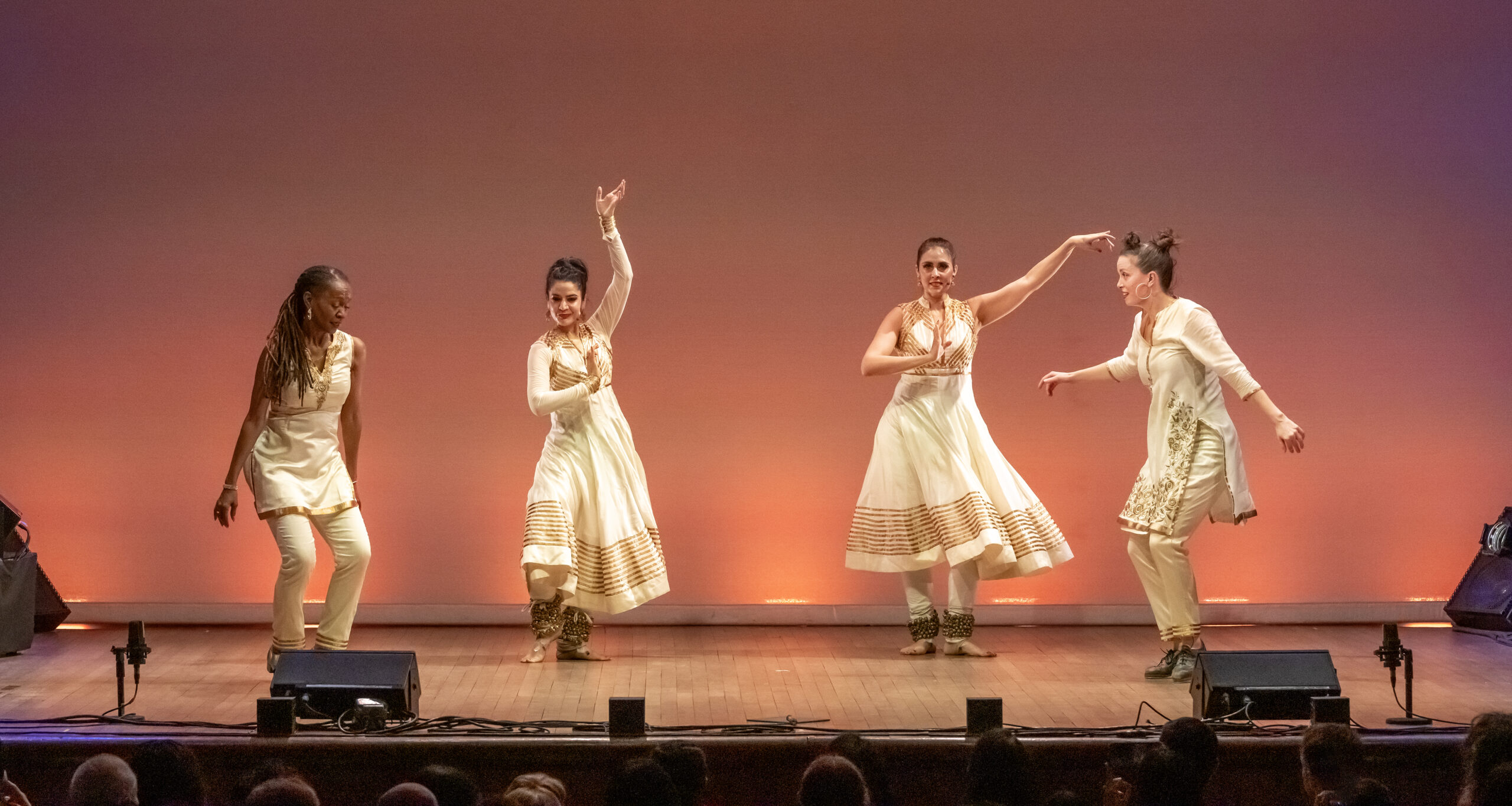 From left to right, dancers Dormeshia, Rachna Nivas, Rukhmani Mehta and Michelle Dorrance. They are in motion. Dormeshia and Dorrance wear white pants, thigh length white tunics, and tap shoes. Nivas and Mehta wear white leggings, long white dresses with golden details on the skirts and bodices. They have bands of bells around their ankles and are barefoot. The tap dancers have a quality of bending and sending energy into the floor. The Kathak dancers are lifted, arms raised, poised.