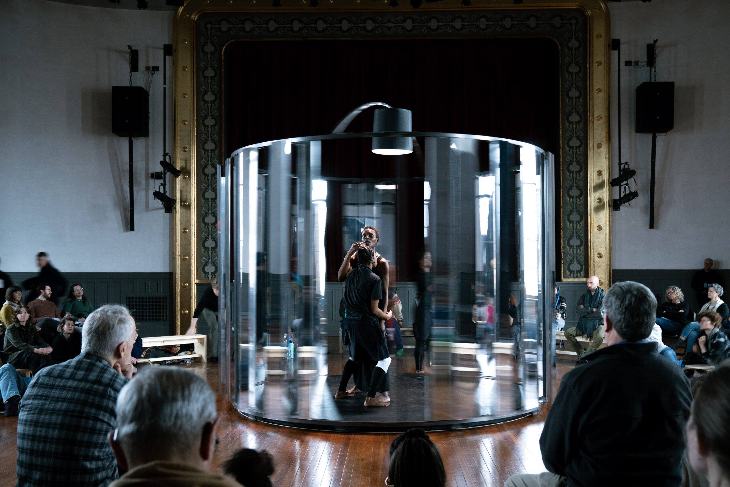 Performers Bria Bacon and Okwui Okpokwasili, both Black women wearing black, stand in the middle of a spinning structure at the center of the room, surrounded by a seated audience. The structure is round with a black bottom and reflective panels about 8 feet tall surrounding it. Through the spaces between the panels, Bacon and Okpokwasili are seen standing close together, facing each other. Becon's knees and arms are bent. Okpokwasili has a hand on Bacon's head and gazes above it.