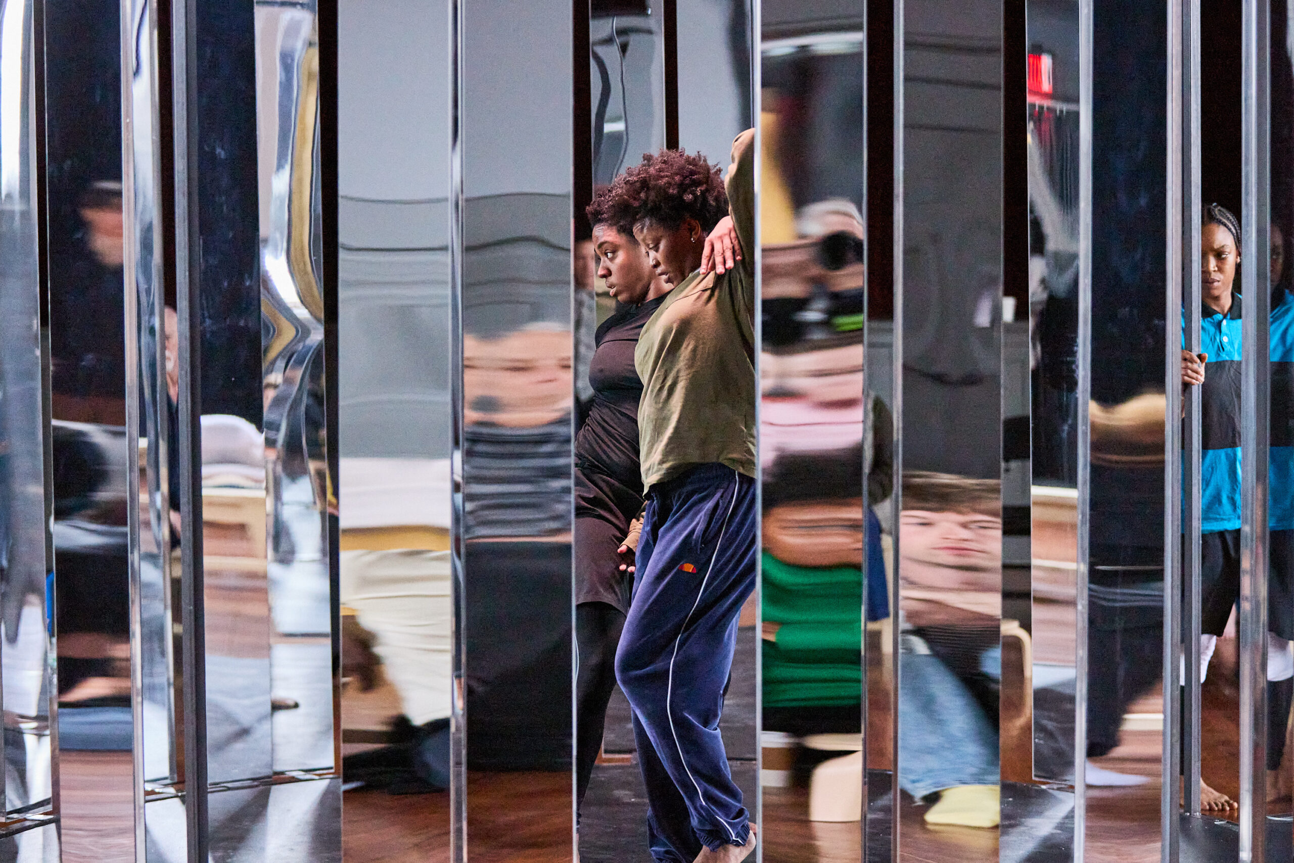 Performers Kris Lee and AJ Wilmore, both Black women, stand inside a metal structure with mirrored panels. Lee wears a black shirt and pants. Her left arm is draped over Wilmore's shoulder, and she looks straight ahead. Wilmore wears an olive-green shirt and navy-blue sweatpants. Her left knee is bent, and her left arm is raised, and she looks down. Bria Bacon, in a blue-and-black striped t-shirt, black leggings, and white knee pads, is in the background, her hand on the structure.
