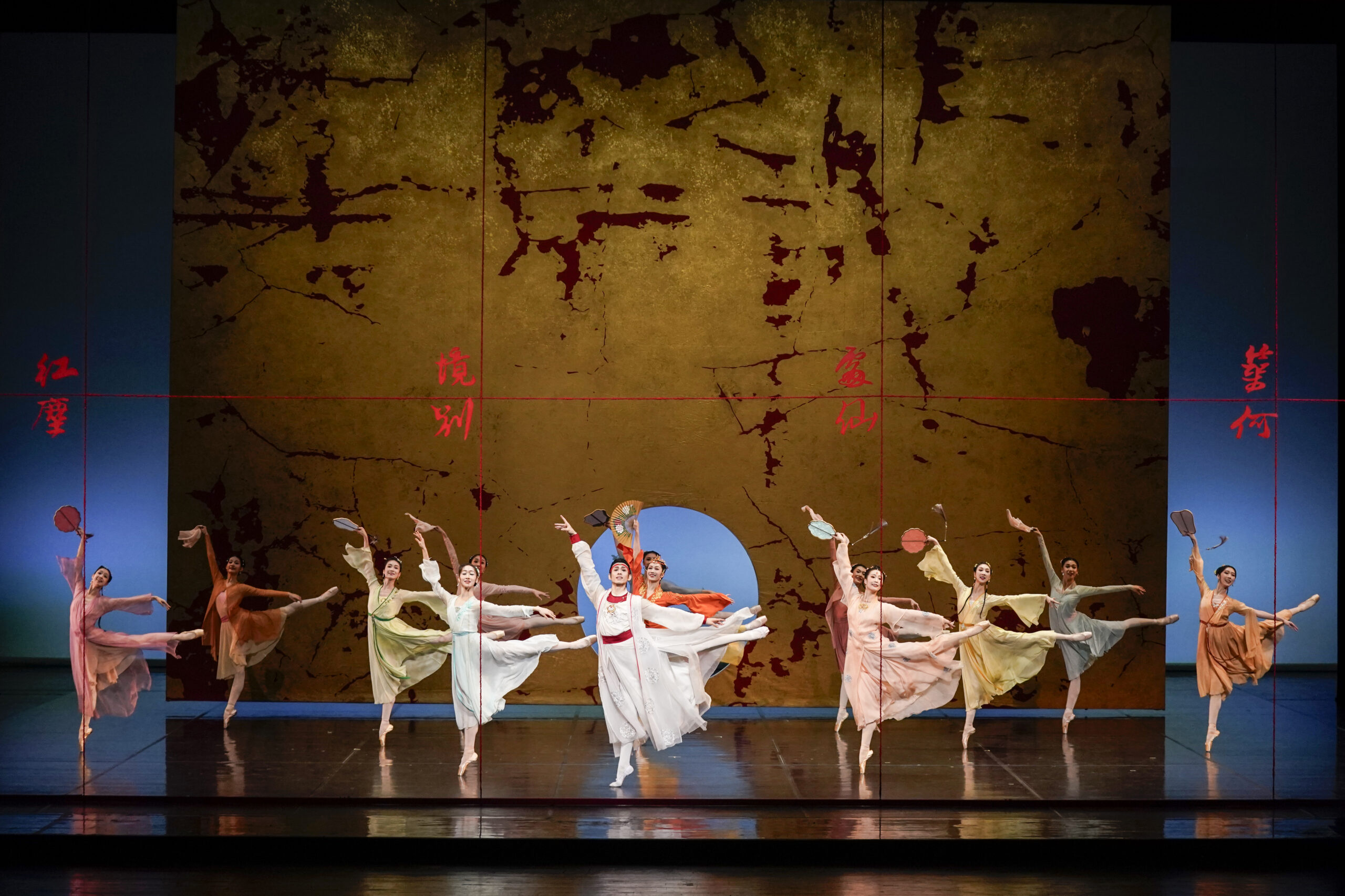 At center stage, Baoyu, dressed in flowing white robes, leads a semicircle of female dancers dressed in pastel, Han-style costumes. The dancers extend one leg in high arabesque-like lines while holding delicate props such as fans and round silk fans, creating a symmetrical and airy composition reminiscent of classical Chinese painting. Behind them is a large golden backdrop textured like aged parchment. Red calligraphic Chinese characters are written across it, associated with the supernatural framework of the story. A circular opening in the center of the backdrop reveals a cool blue background, evoking the moon or an otherworldly portal. The stage lighting highlights the dancers’ flowing sleeves and soft colors—peach, pale green, yellow, and ivory—suggesting the youthful elegance of the family's girls.