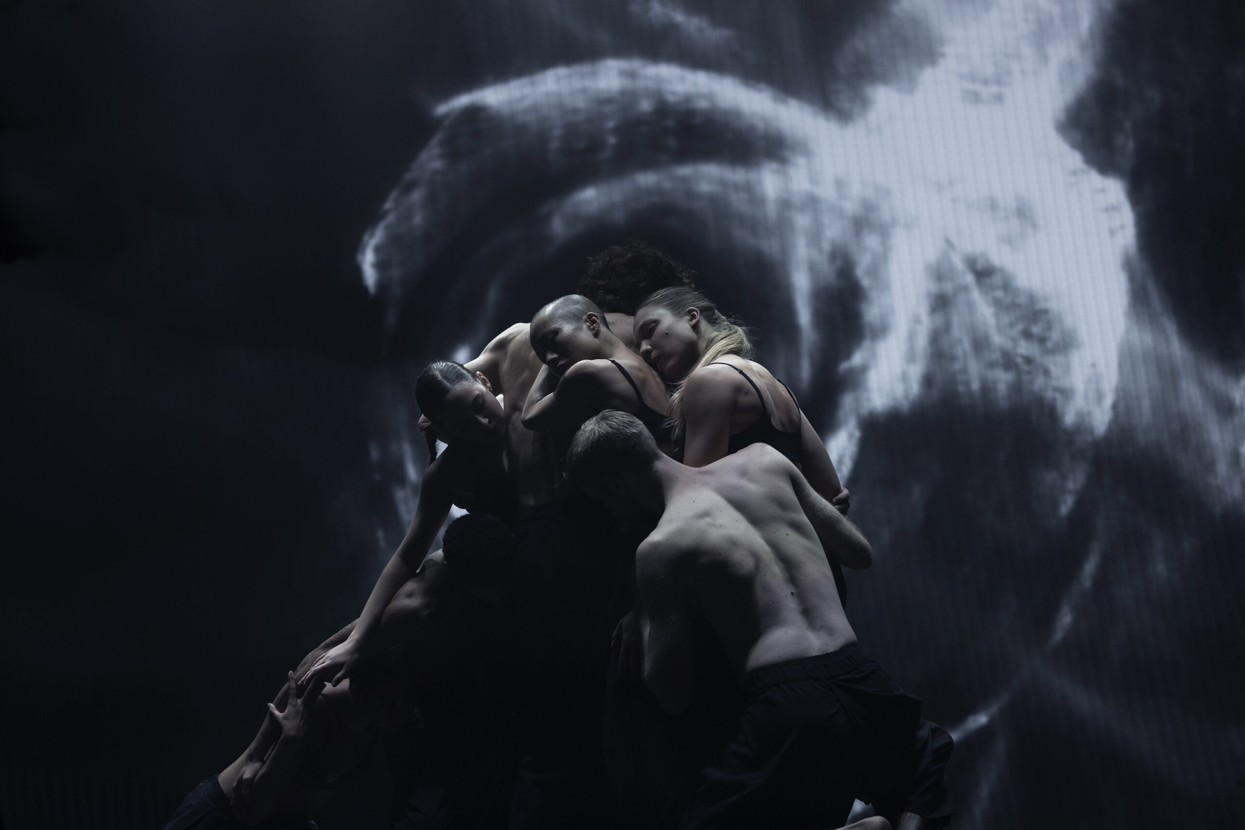 A group of dancers stands in a tight cluster, in shadowy lighting. A black and white video projection lights up the backdrop behind them.