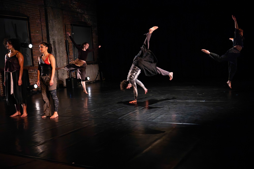 On a black marley floor and dimly lit stage, Amalia Colon-Nava and Anna Scattoni stand far left facing the audience. Behind them, three more dancers are captured in motion. Amanda Rattigan and Kayliani Sood are leaping, as Ian “Seven” Tackes is mid-handstand.