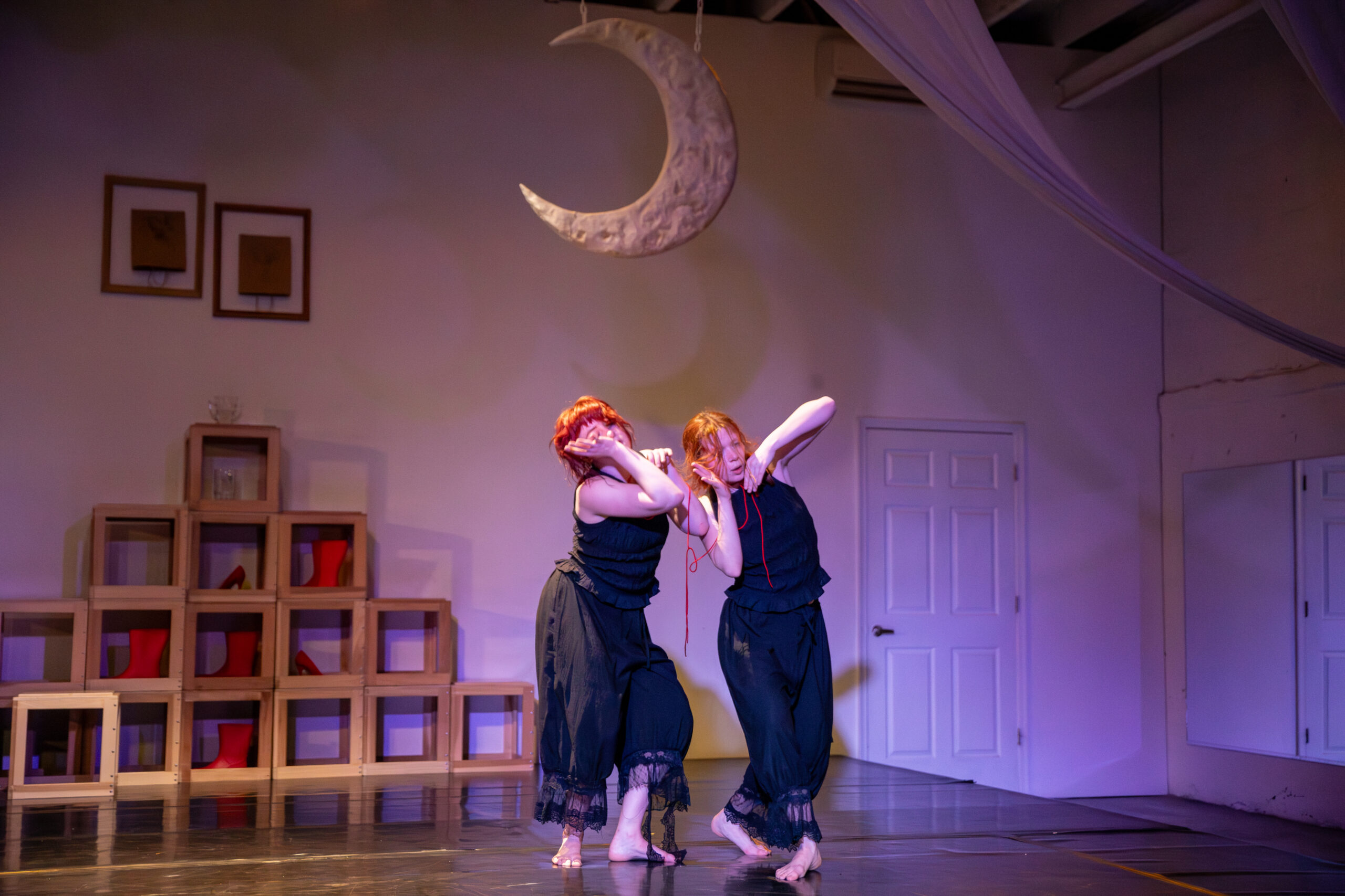 Two white women with bright red hair pulled back loosely, wear black pants and tank tops and accentuate the curves of their waists, leaning into their hips and slightly covering their eyes with elbows bent at different angles. They are loosely connected by a thin, red thread and in the background there is a hill constructed of wooden blocks against a white wall. Completing the scene are red galoshes, two picture frames hung above the hill and a large new moon hung from the ceiling.