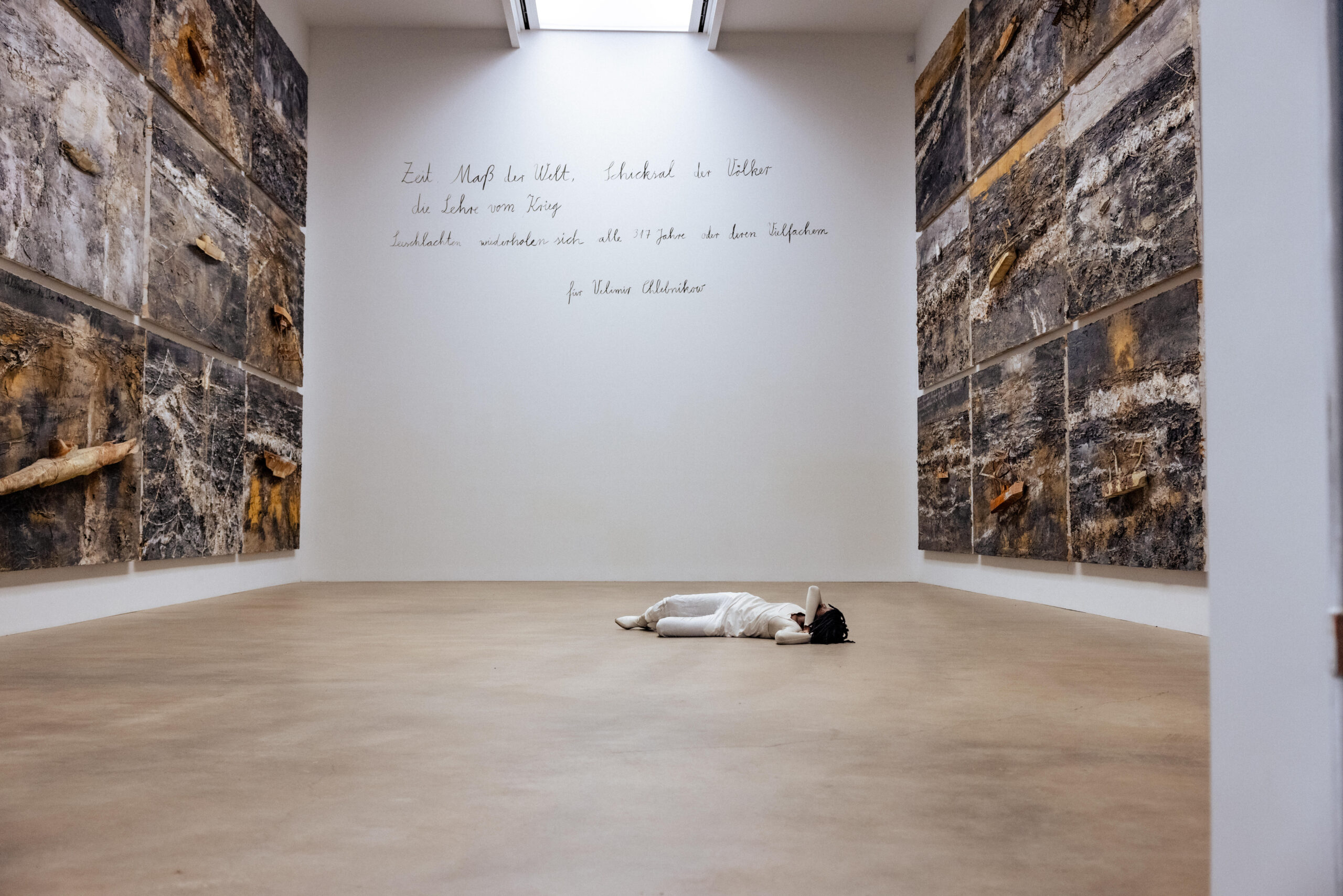 Dressed in white, performer Marguerite Hemmings lies prone on the floor of a large museum gallery at Mass MoCA between monumental earth-textured wall panels, beneath handwritten text on the far wall.