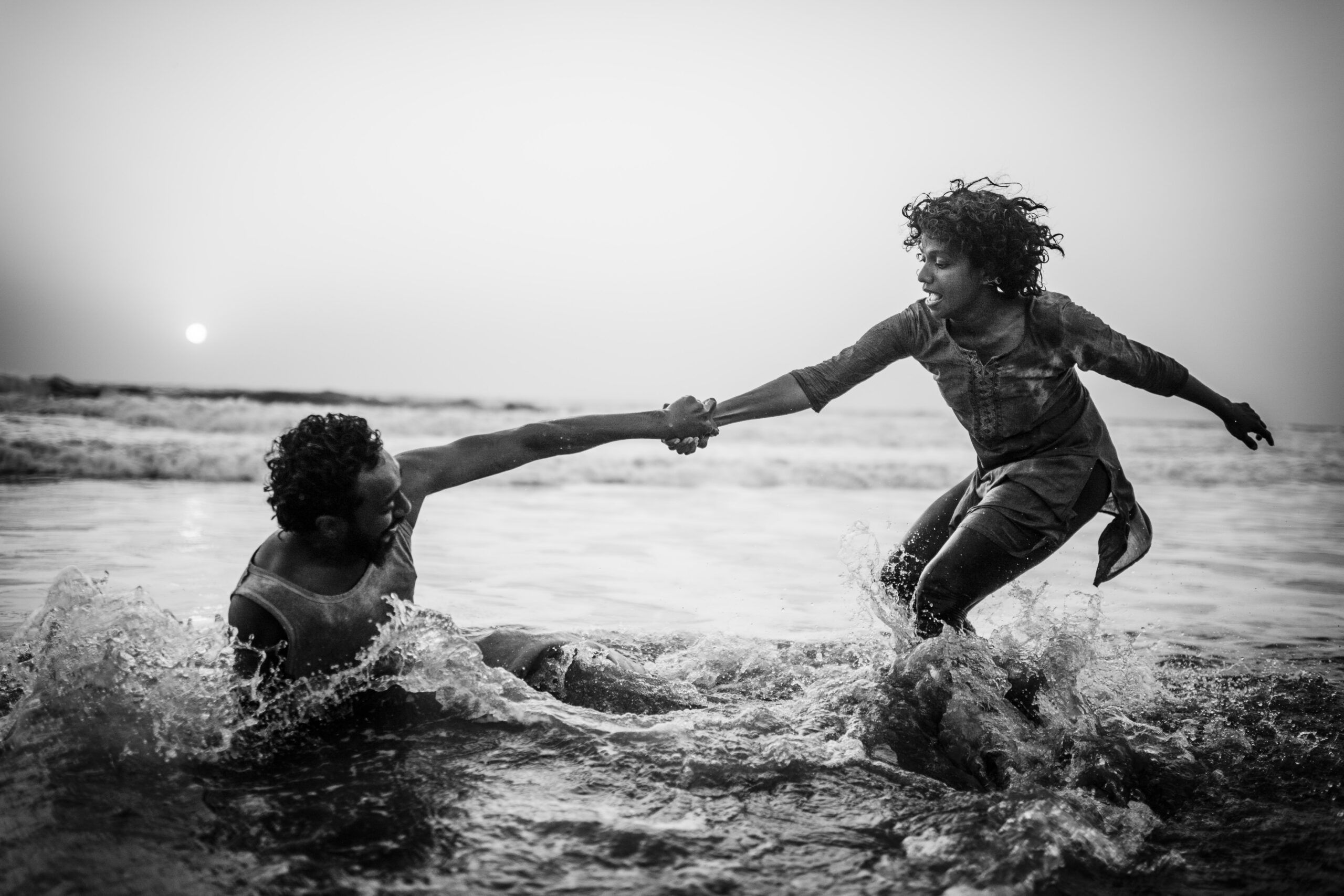 Two dancers practice the Contact Improvisation principle of weighing-out while in the shallows of the Arabian Sea at sunset. Small waves roll and crash in the background. The sun is a hazy dot in the upper left hand corner. Water crashes around the pair–a woman with short bouncing curls who stands, knees bent, and a man with short dark hair who presses one hand down into the water and sits on his side. Perhaps the wave has taken him out. The woman extends her left arm for balance as she reaches her right toward her partner. The two hold hands at the wrist as the water splashes around them. The photo is in black and white.