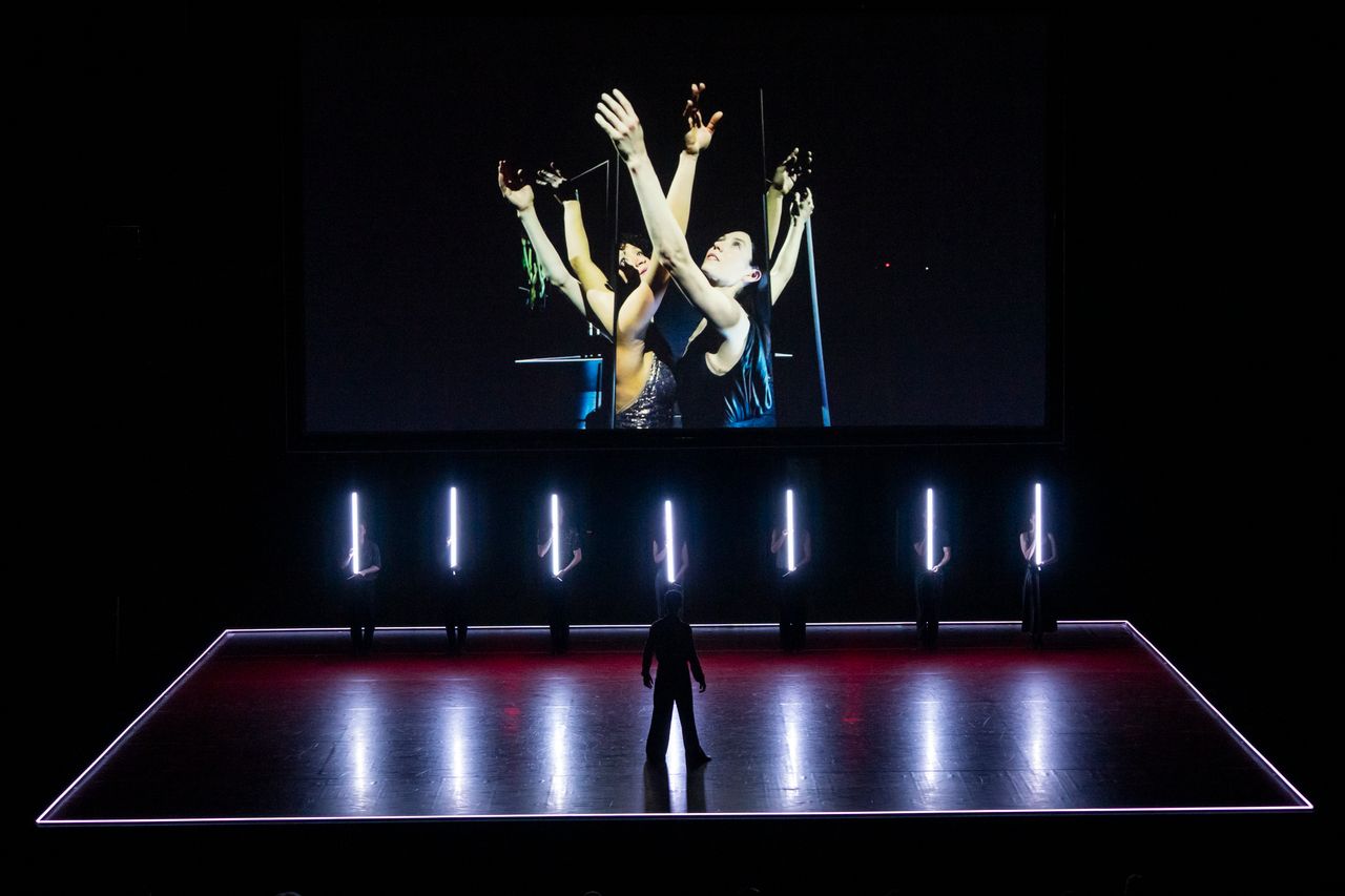 On a screen, Rachel Hutsell and Daphne Fernberger stand close together and reach up, crossing arms. The pair stands between a set of mirrors so their image reflects, causing the illusion of three sets of arms instead of one. On the stage below the screen, seven L.A. Dance Project ensemble dancers stand at attention, each holding a long, thin tube light vertically in front of them. At the front of the stage, a dancer in silhouette stands in a strong stance, feet wide, watching the scene unfold.