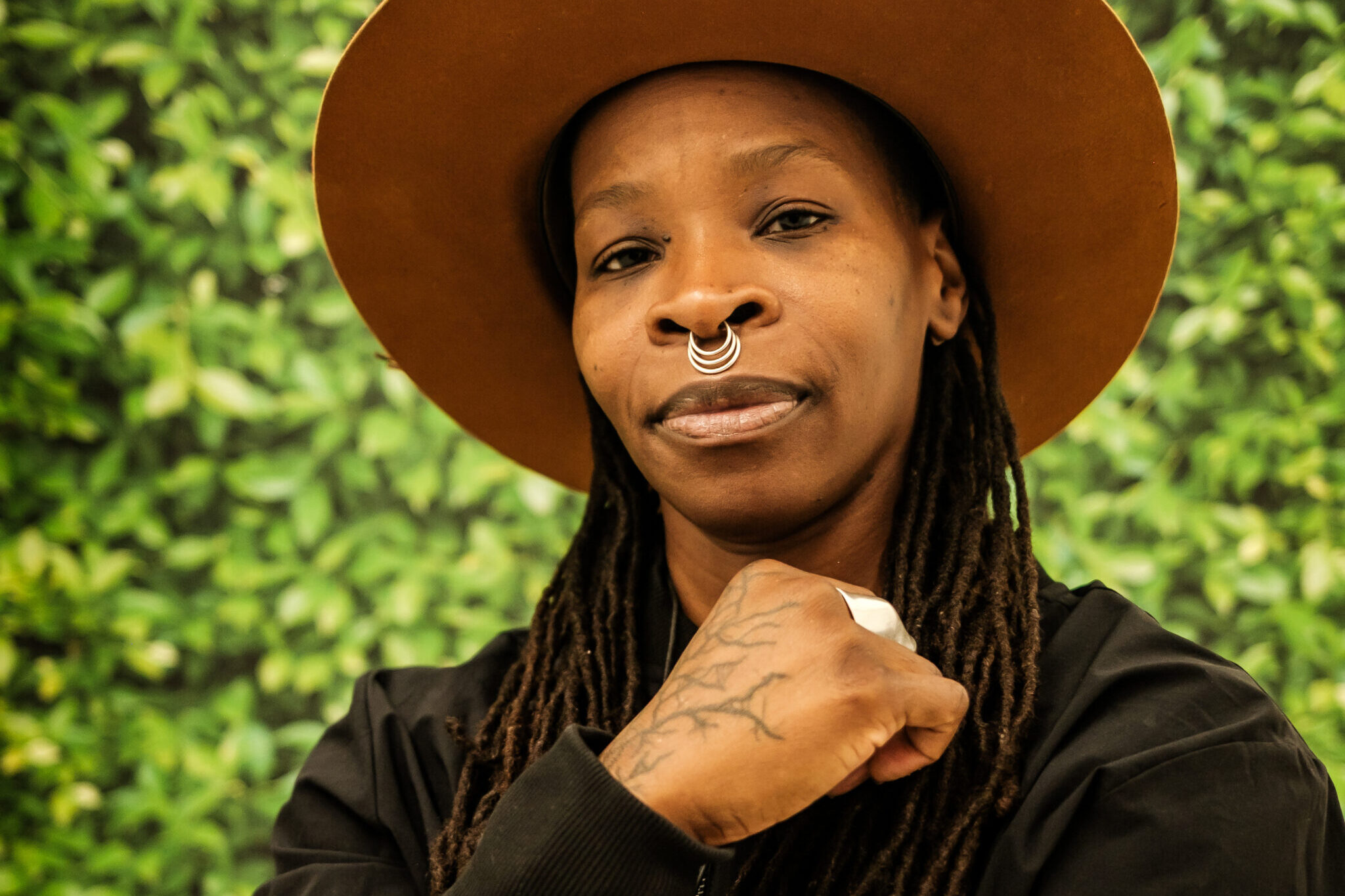 Sage Ni'Ja Whitson, a queer transgender Black person with long black-brown locs faces the camera with a soft, powerful smile. They wear a round-brimmed brown hat, a triple-ringed silver septum piercing, and a black jacket. One hand with vein-like tattoos is held in a loose fist near their sternum, while the other is wrapped loosely around their stomach. They stand in front of a leafy green backdrop.