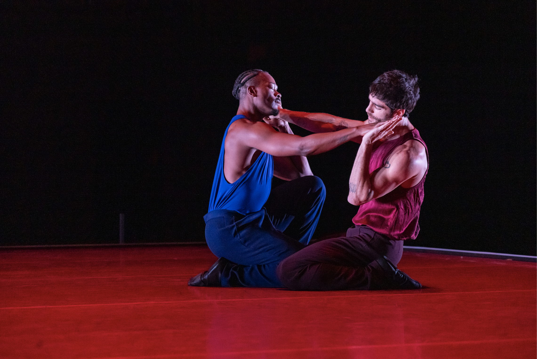 David Adrian Freeland Jr., wearing a blue sleeveless top and pants, and Morgan Lugo, wearing a red sleeveless top and pants, kneel facing each other on the red-lit stage. With closed eyes and tilted heads, they touch palms, one arm straight and the other bent by their cheeks.