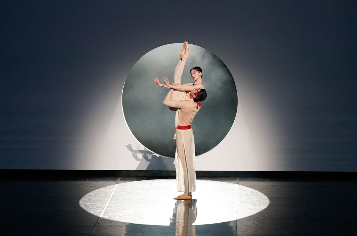 The image captures a romantic and dreamlike moment between Baoyu and Daiyu. In front of the white, elegant backdrop, Baoyu stands grounded at center stage, gently lifting Daiyu high above him. Fully embraced and supported by Baoyu, Daiyu extends both legs vertically in a striking display of control and flexibility and her arms curve softly in a refined, sculptural pose. The moon gate at the backdrop echoes the circular spotlight on the floor, framing the dancers’ pale, flowing costumes with subtle red accents and adding a quiet intensity. The composition feels balanced and intimate, capturing a suspended, dreamlike moment of elegance, control, and emotional restraint.