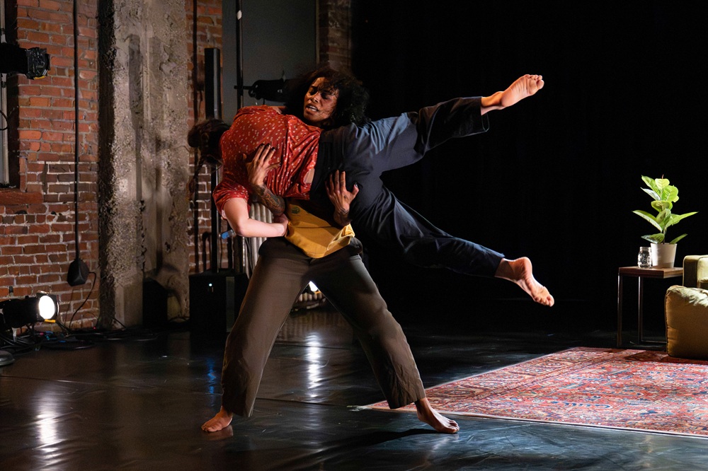 Chloe Marie and Tamar Gutherz are captured on stage mid-stunt in this action shot. Chloe is standing upright grasping onto Tamar who is horizontal in their arms with legs extended long. There is an area rug and an end-table with a potted plant in the background.