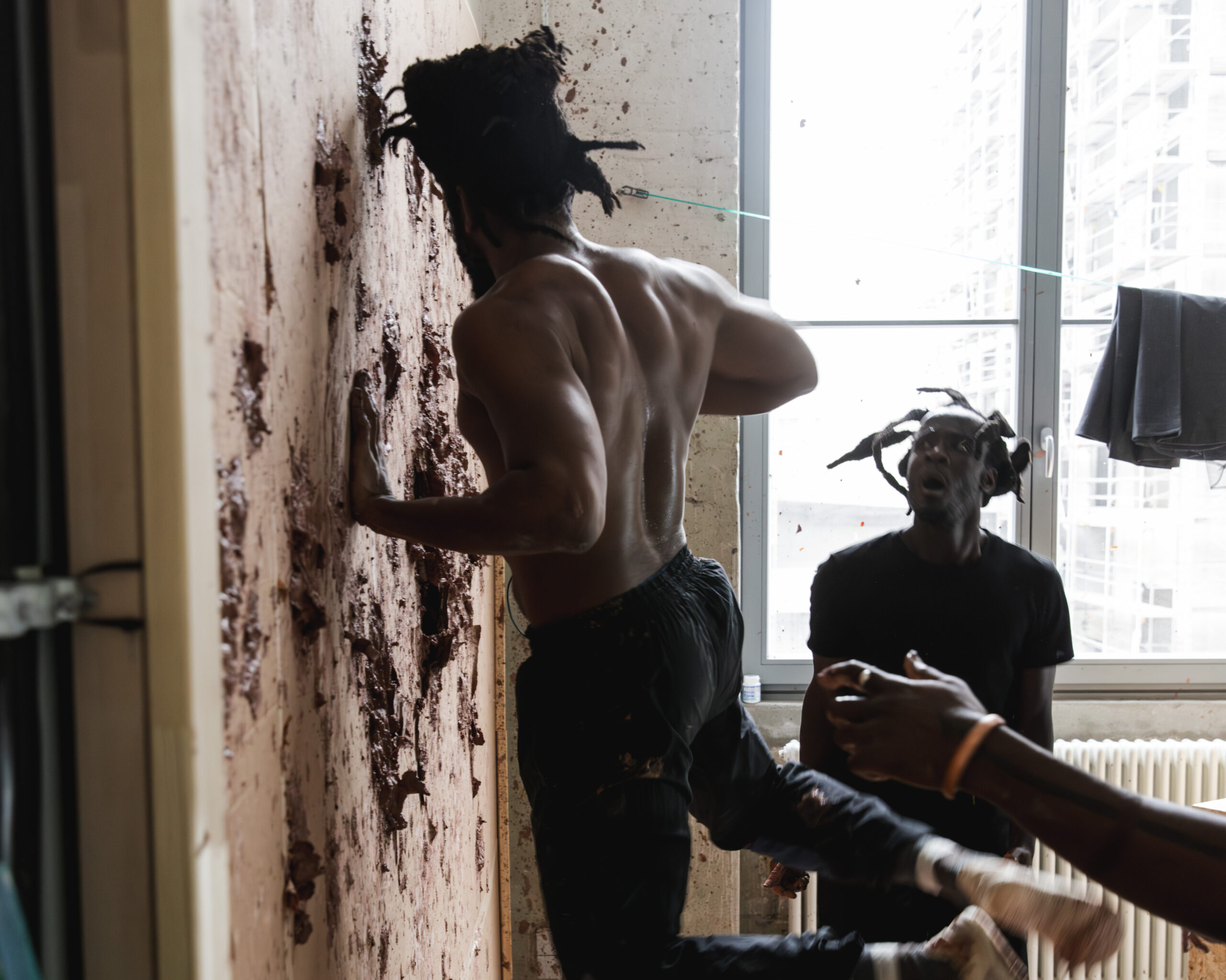 In a scene from Dambudzo, tyroneisaacstuart punches red clay into a cardboard wall, face turned away from the camera, as Tatenda Chabarwa looks on at this scene, face aghast in horror. Both are Black men outfitted in plain black clothing that both contrasts their dark brown, Black skin tone and captures remnants of red clay that spatters off the wall.