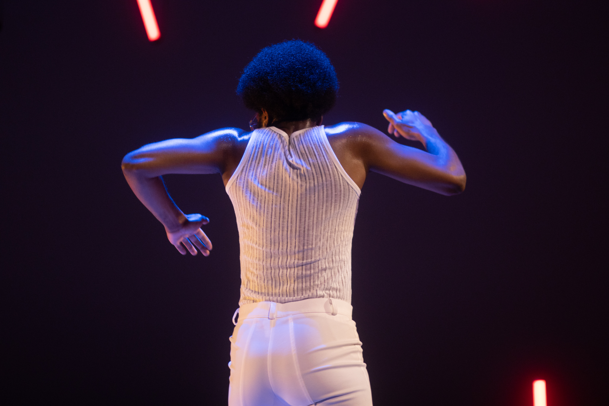 One figure with textured hair and deep brown skin lifts their elbows to shoulder height, their back to the camera, wearing all white.