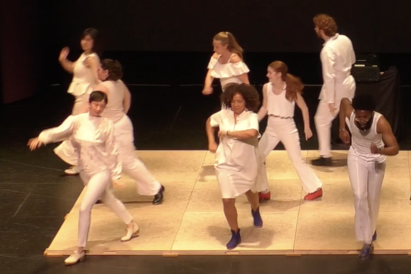 Thirteen dancers in all white and different color tap shoes dance joyfully off of wooden boards in all directions. Their bodies blurred in space.