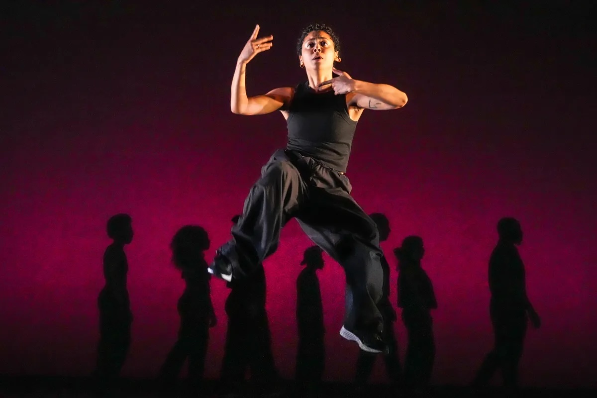 One dancer looks straight out into the distance, their face pinched in a look of concern, hands reaching back towards their chest. They are jumping up past a lineup of silhouettes imprinted on a blood red background.