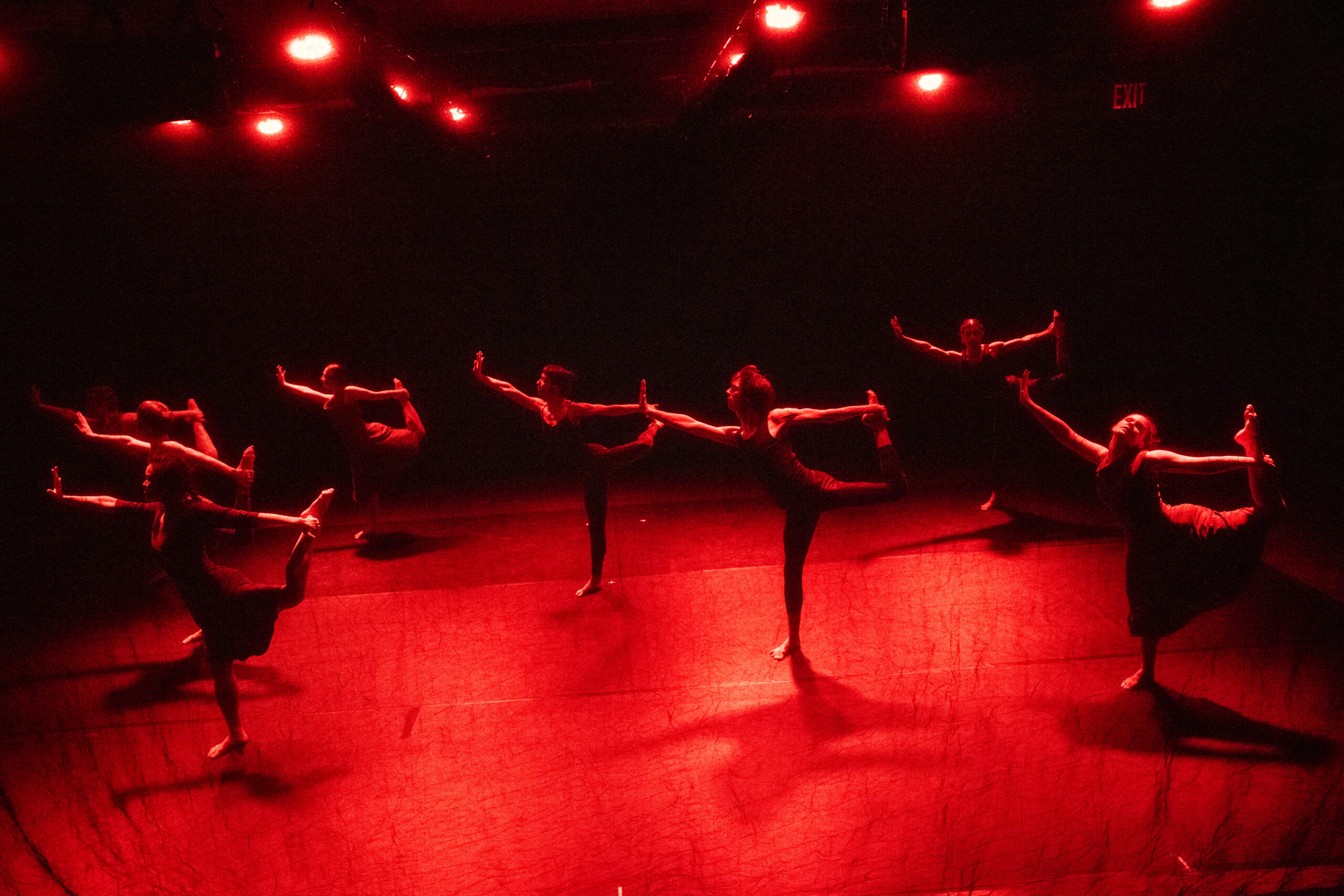 Six young dancers stand in profile, all facing right, under bloodred stagelights. They balance on their right foot, while holding their bent left leg with their left hand behind them. Their right arms are extended in front of them, their palms flexed, as if threy are saying "stop."