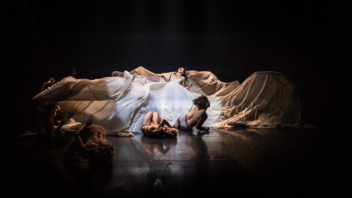 The vast white skirt of a female dancer spreads out across the center of the stage, drawn and lifted by dancers concealed beneath it, resembling a giant wave. The dancers are constantly struggling to crawl out from within this undulating mass of soft fabric. With their upper bodies bare, they curl up on the ground, suspended in a state between weightlessness and struggle. The spotlight focuses on the white fabric and the figures at the center, plunging the surrounding space into darkness.