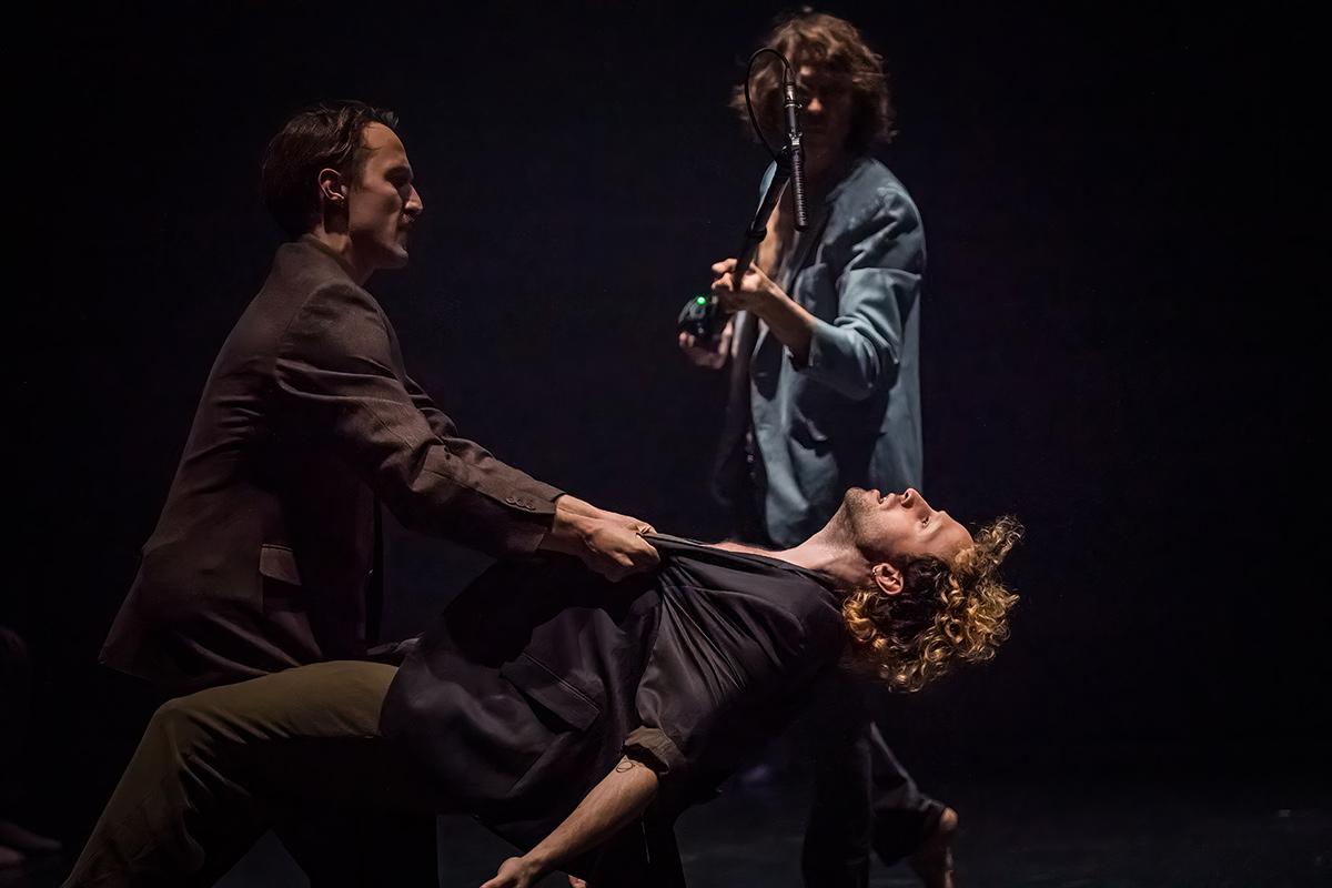 One dancer grabs the other by the collar, suspending him in midair. The lifted dancer’s head tilts back and his chest opens, embodying a forced exposure and a sense of loss of control. The two dancers appear to have just been in a struggle. Behind them, a performer holding a microphone stands in the light, as if witnessing or intervening in this moment.