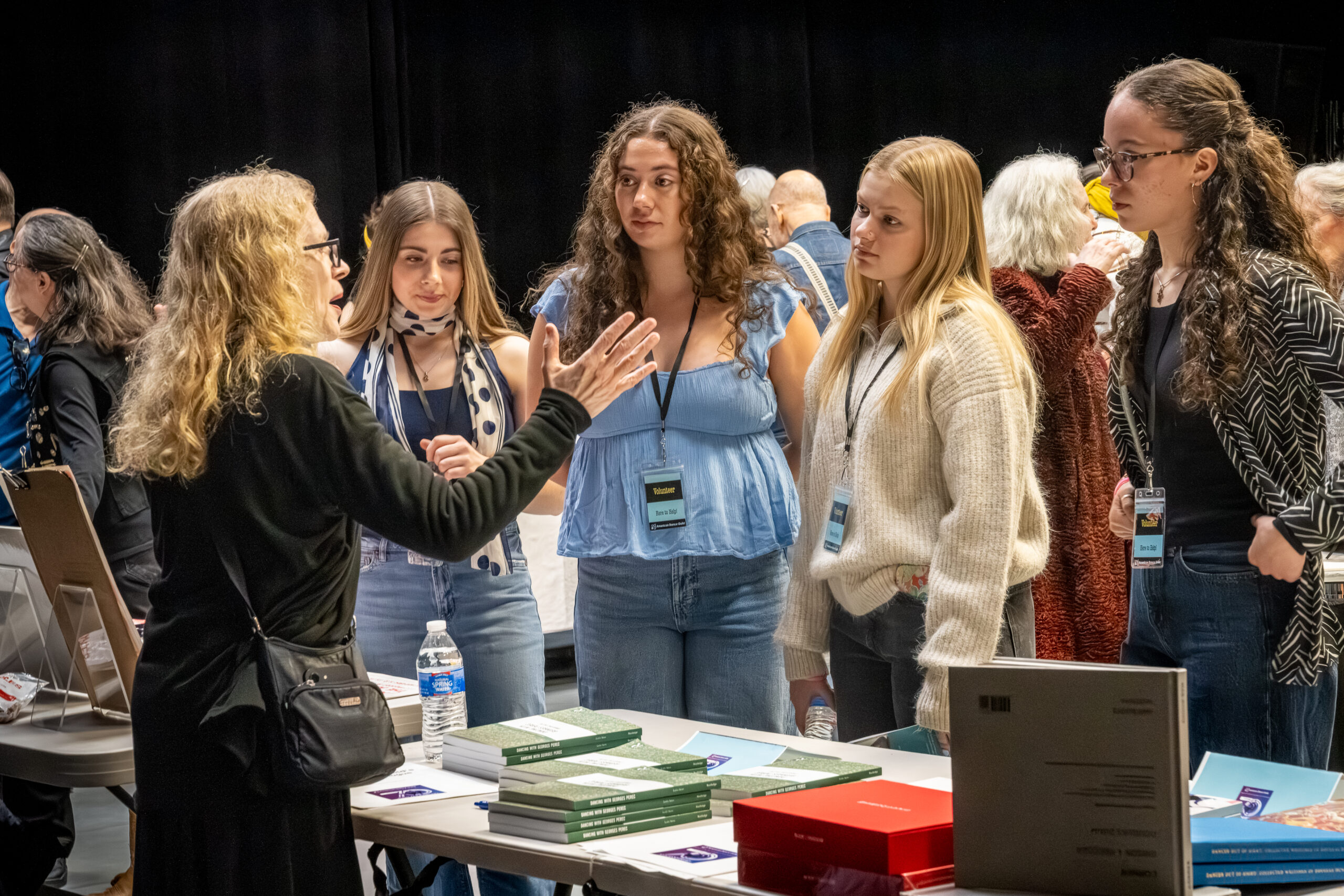 Dancer and Author Leslie Satin stands behind her book table, stacks of green spines in front of her. She has long strawberry blonde hair and long black sleeves. She is gesturing with her right arm up and palm wide open while she speaks to a group of four young women.