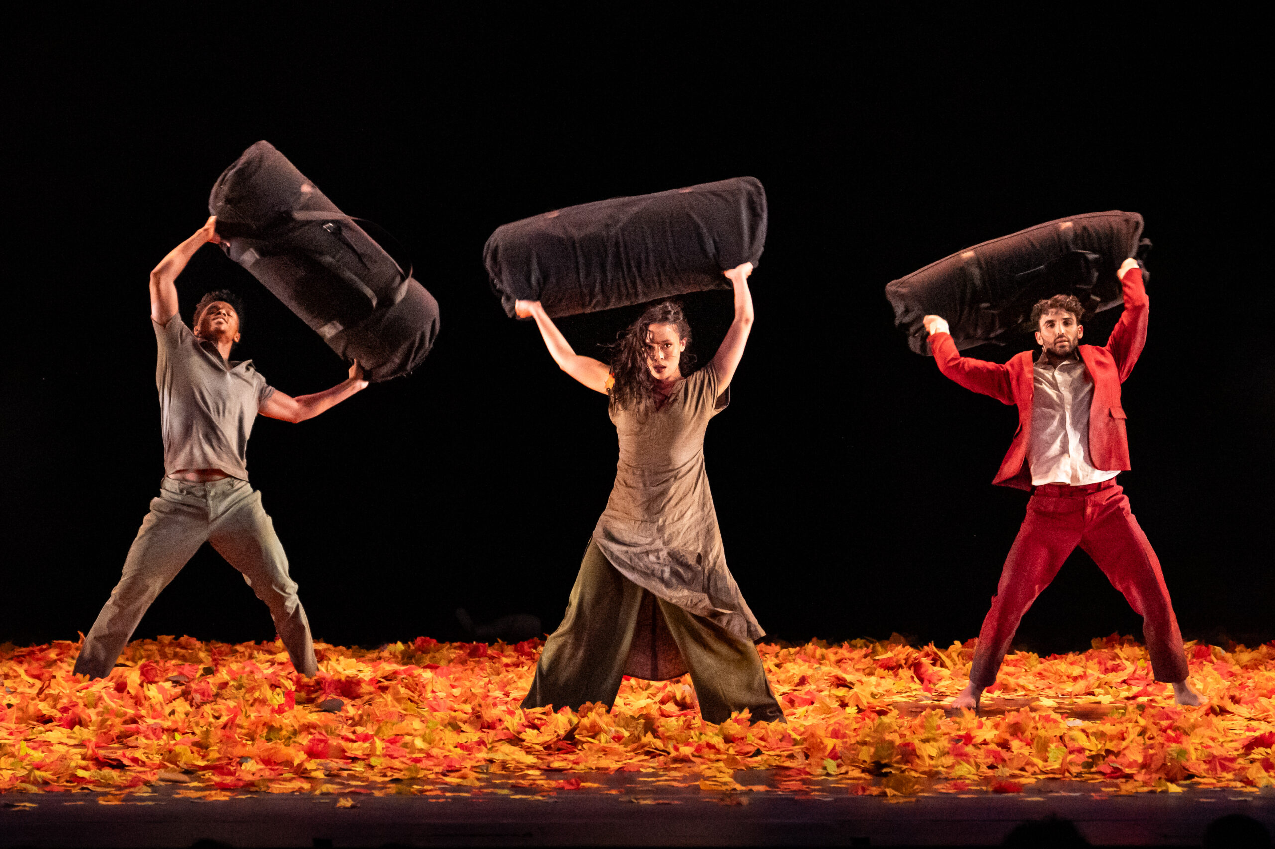 Three dancers, two men and one woman, stand on a stage covered in bright autumn leaves. The background is black. They stand in a wide stance, holding thick black rolls over their heads. The man on the left, in gray pants and a t-shirt, looks up at the roll. The brunette woman wearing green pants and a brown tunic stares directly out. The man on the right, dressed in a red suit and white dress shirt, also looks straight forward.