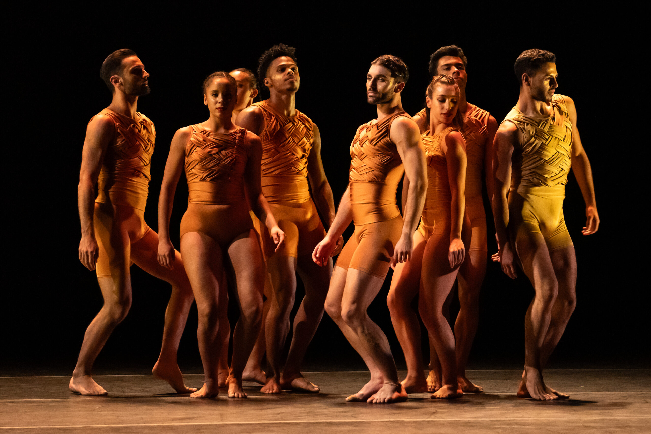 Eight dancers, three women and five men, stand in a group with their knees slightly bent, facing different directions. They wear tight shiny orange-gold tops and shorts. Some look over their shoulders, and some look straight ahead.
