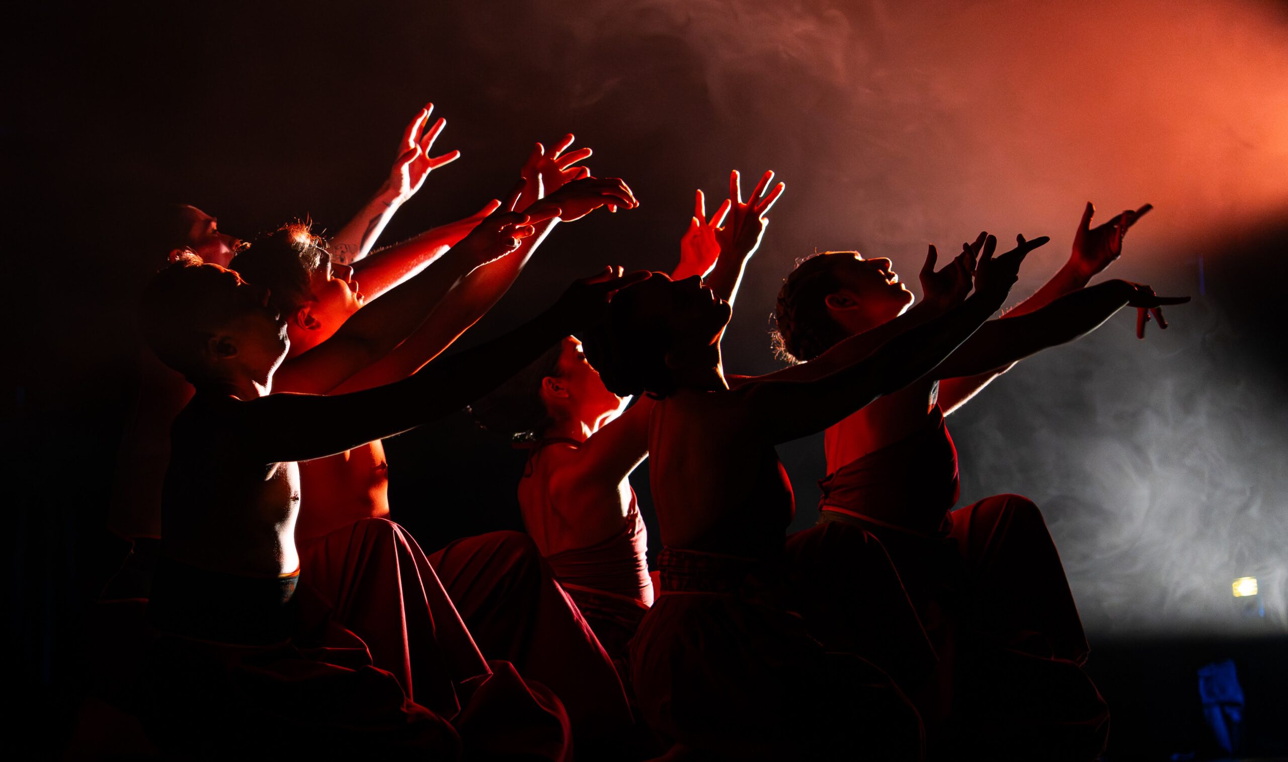 A group of shadowy-figured dancers in red kneel in profile. They reach their fingers toward a moody, red light in the upper right hand corner. The whole scene is bathed in haze