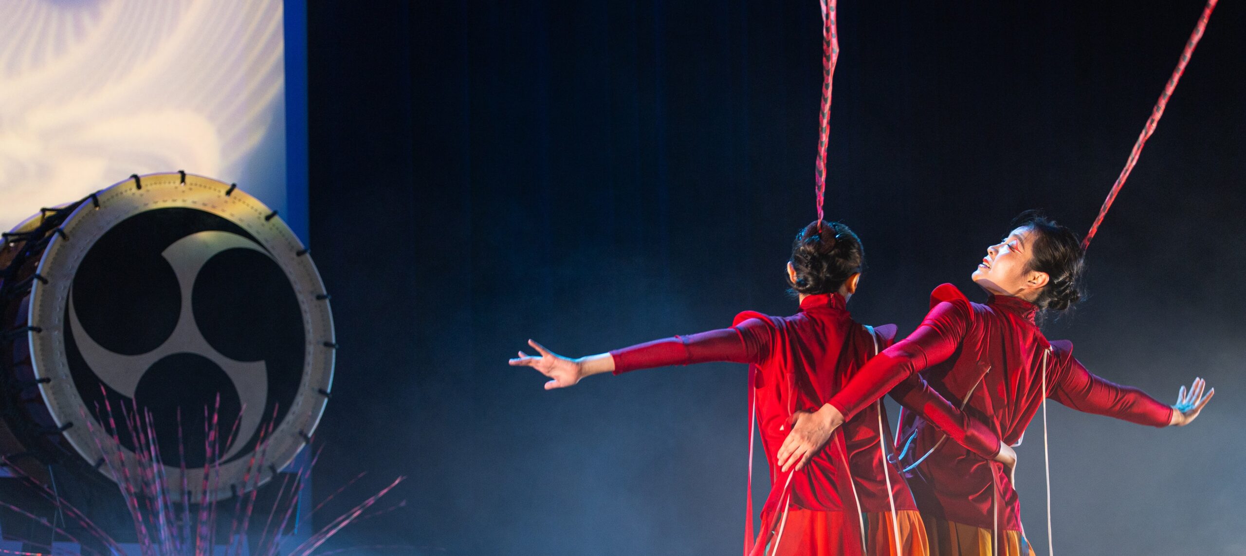 Two dancers in long-sleeved red tops face away from us with arms round one another’s waists as their free arms reach outwards. There are singular, red feathers extending from their heads many feet into the upwards space. To the left of the duet, we see a large Taiko drum.