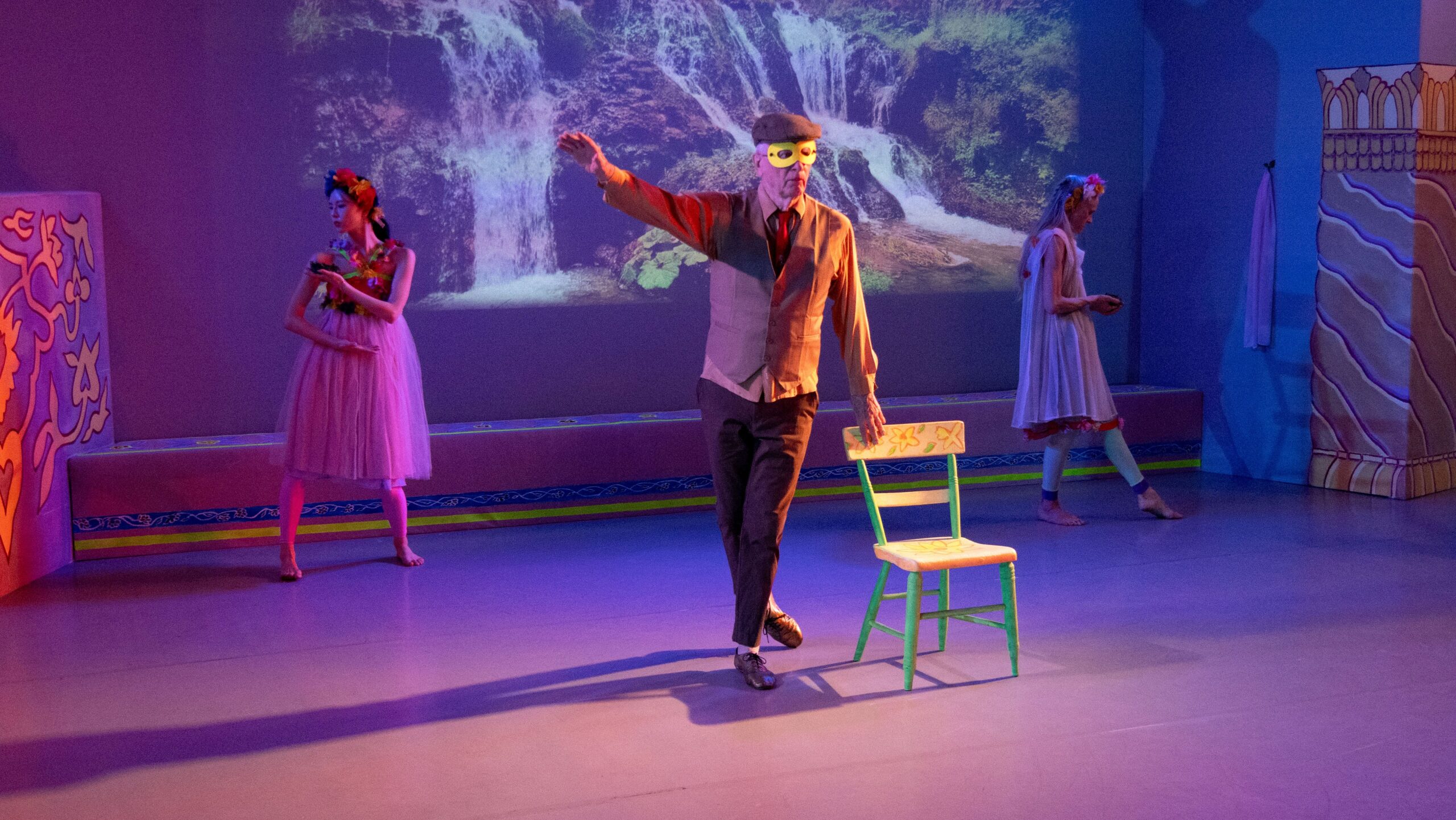 Douglas Dunn stands wearing a bright yellow mask which covers his eyes. His right arm is extended to his side while his other rests on a wooden chair painted with yellow flowers. He wears a grey vest, red tie, and dark pants--a contrast to dancers Dongri Suh and Janet Charleston who stand behind him weaering flowered garlands around their heads and wear tulle skirts. A video of two waterfalls is projected onto the wall behind them.