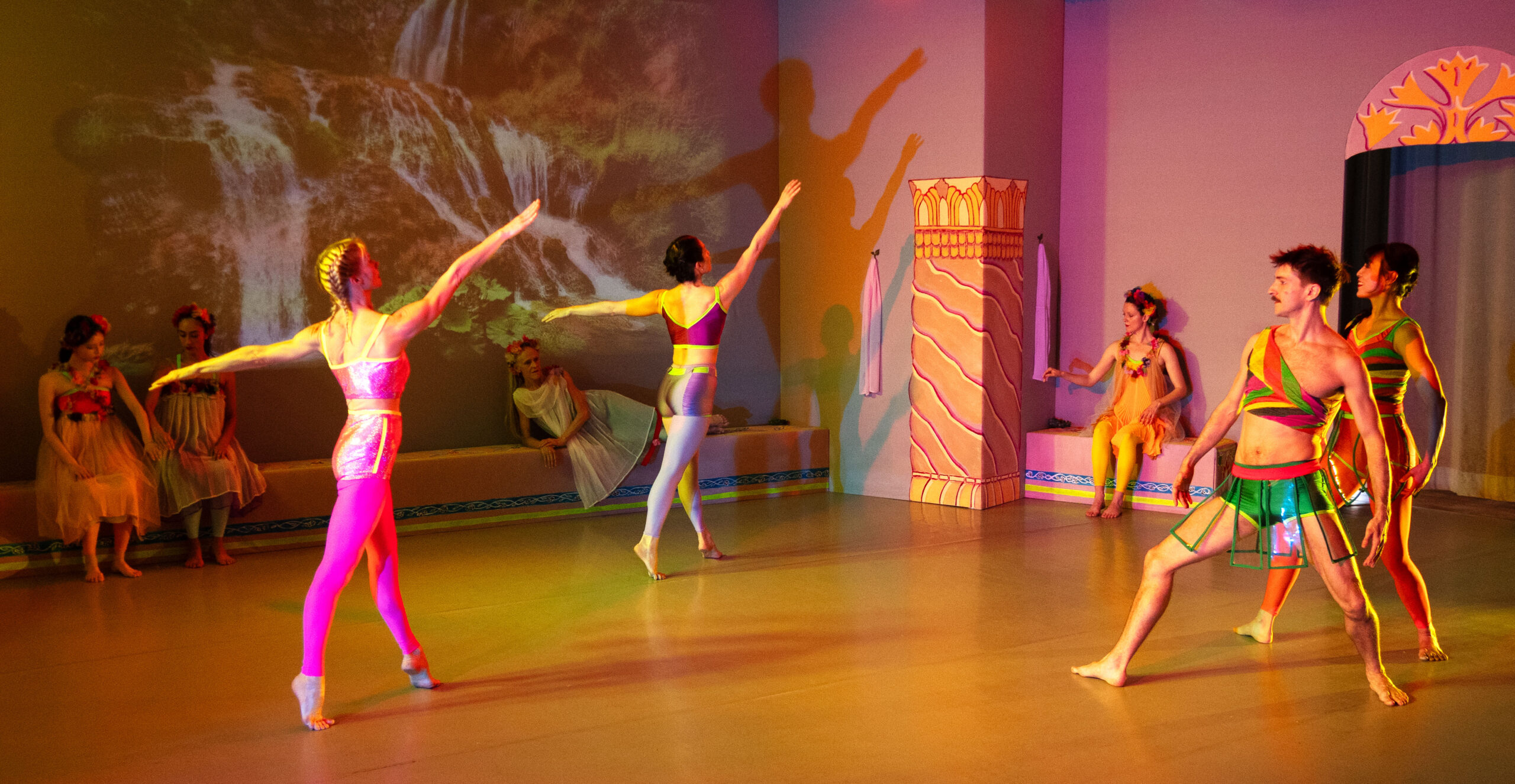 Eight dancers pose within a brightly-lit room, each wearing colorful costumes and with painted pillars around them evoking ancient Greece. Four of them sit or recline along the wall upstage, wearing flowers in their hair and adorned in tulle skirts. In front of them are two women in profile, balanced on the balls of their feet and extending their right arms as they look up. Facing them and to the right, two more dancers stand with their bent thighs wide apart, their curved arms away from their sides like warriors. On the wall upstage of all of them is a tranquil video image of two waterfalls.