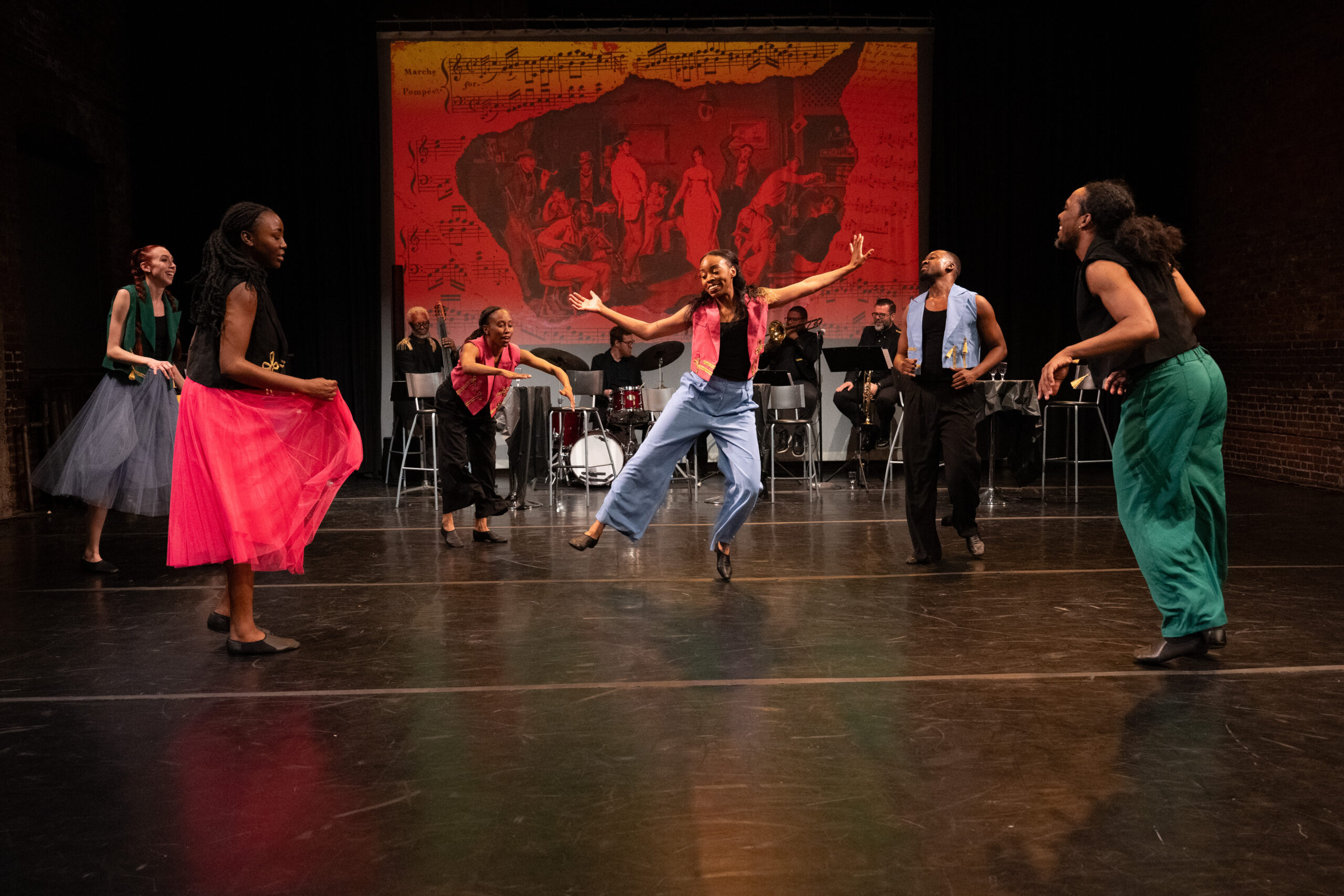 A group of five dancers, three women and two men, form a circle around a female soloist. The soloist, wearing a vibrant pink vest over a black top paired with light blue, wide-legged pants, moves exuberantly with her arms out akimbo while standing on her left toes with her right leg out to the side. A live five piece jazz band, including a piano, drums, a bass trumpet, and trombone, is visible behind the dancers upstage. A projection on the brick wall in the back displays a collage of sheet music and colonial artwork of a scene from a pub.