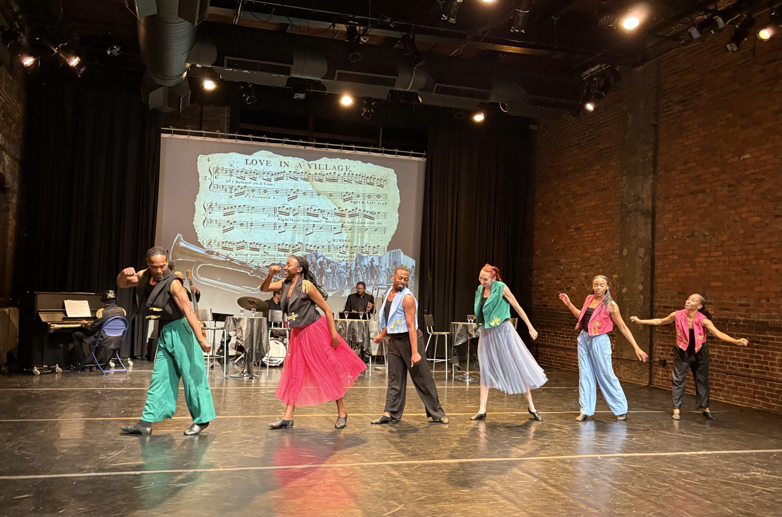 A group of six performers, four women and two men, are in a horizontal line dancing across the stage. They are dressed in colorful outfits that include pink skirts and colonial military styled vests in blue, pink, green, and black. Projected on the way is a background featuring a projection of sheet music titled "Love in a Village." A live five piece jazz band, including a piano, drums, a bass trumpet, and trombone, is visible behind the dancers upstage. The venue itself has a brick wall and dramatic lighting that aid in creating an engaging atmosphere.