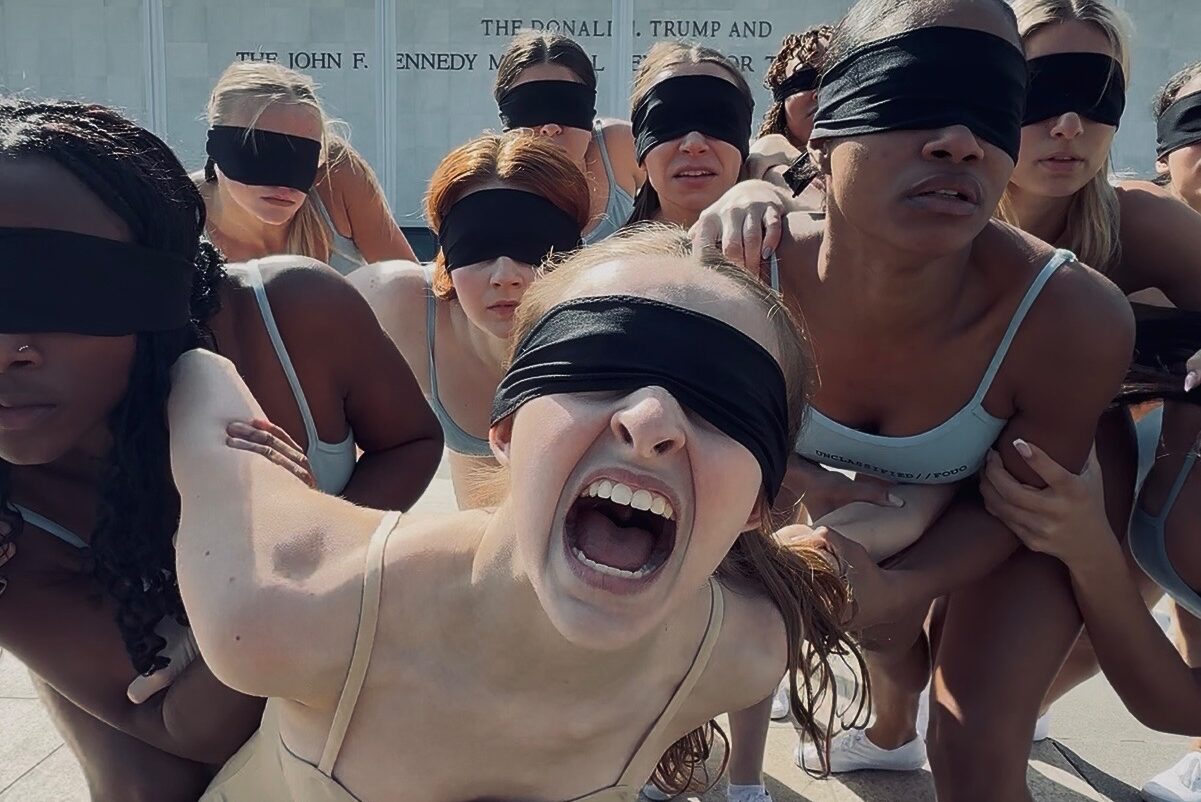 In a close up photograph, ten dancers in spaghetti-strap leotards lean in, their eyes covered by a sheer black cloth. The middle dancer, closest to the camera, is mid-scream. Behind the dancer is the newly engraved building signage reading “The Donald J. Trump and John F. Kennedy Center.” Some words are covered by the dancers’ heads.