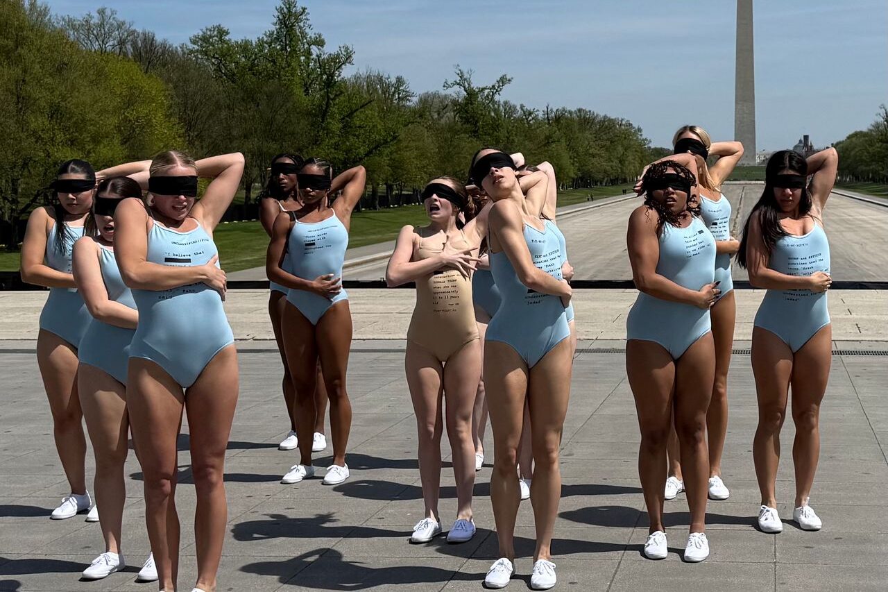 An ensemble of dancers stand upright wearing form-fitting leotards and lace-up white shoes. All have a sheer black cloth covering their eyes. Their left arm reaches behind their heads, elbows flexed, and their right arm snakes across various places of their waist, ribcage, and back. All of the dancers look in varying states of unease.