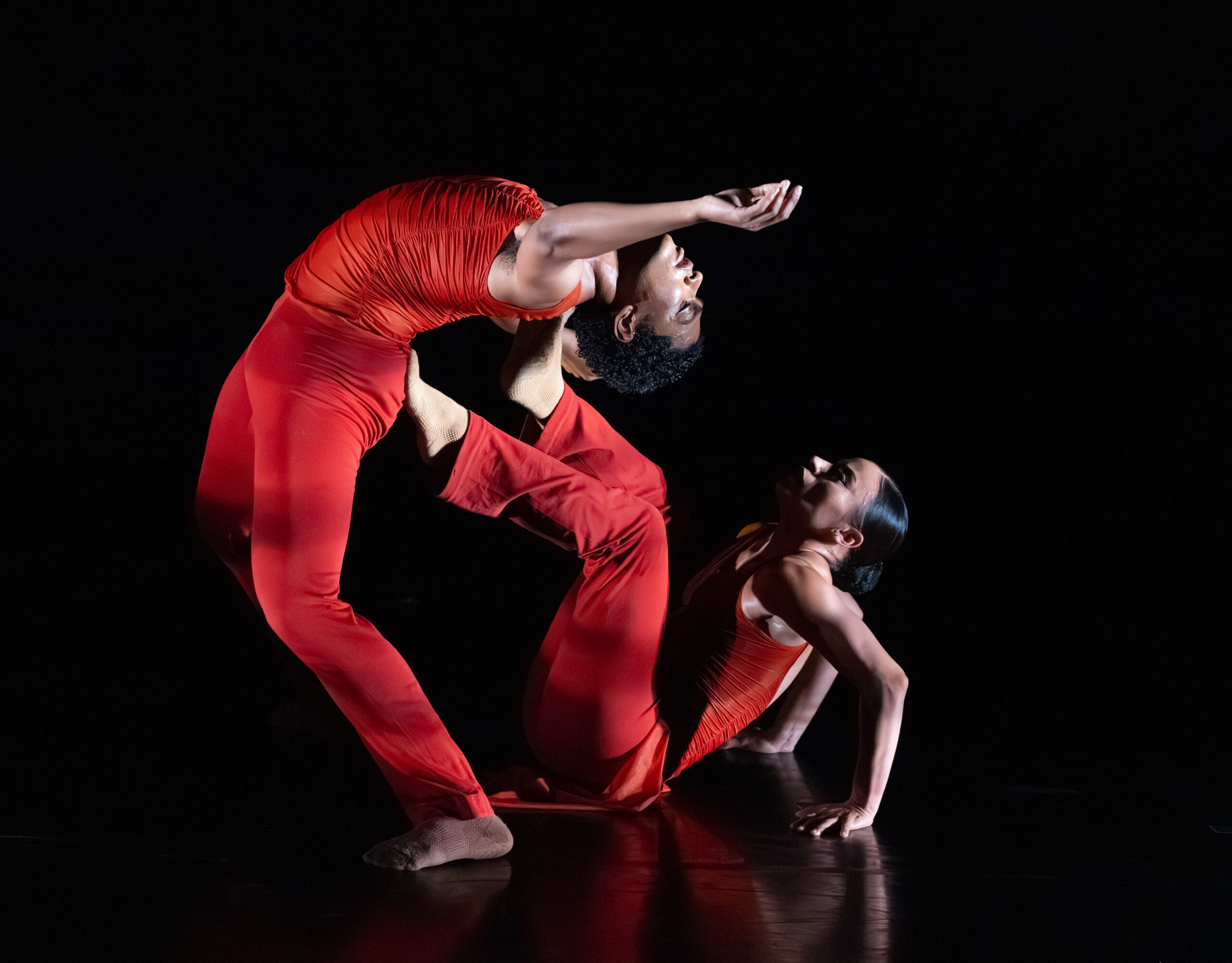 Two bodies are centered in the image, their brown skin a vibrant complement to the red skin tight garments they wear. One dancer balances on her coccyx bone and hands while supporting a tall figure lushly leaning against the seated dancer's two feet.