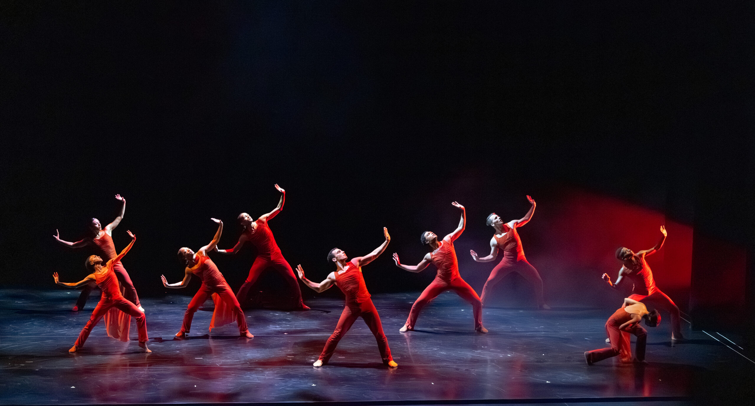 9 dancers dressed in red spread across the stage. 8 of these dancers’ arms part above their heads, eyes tilting up as if to look towards the heavens hoping for strength. One dancer is an exception, keeping their head down to the ground while in a shallow diagonal lunge.