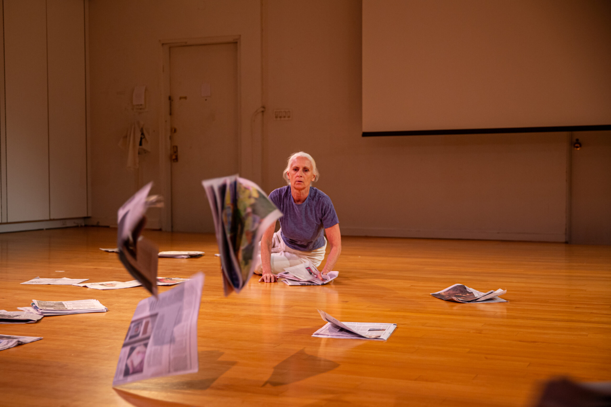 Dancer KJ Holmes, leans onto her left hip, legs folded behind her, and both hands planted on the hard wood floor. She wears a blue t-shirt, white pants, and her grey hair is pulled back from her face. Newspapers are scattered about the floor around her, and she watches the pieces she has just thrown at the camera as they fly away from her. They are blurred by their motion and closeness to the camera.