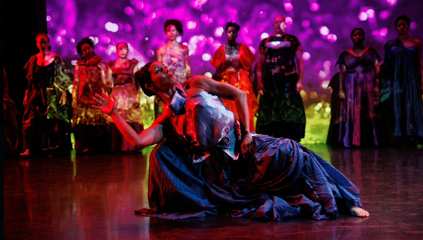 A dancer dips into a low second position, leaning over her right knee, chest and chin lifted, an open right hand reaching. Her silver top and the billowing folds of her skirt shimmer in the blueish light. Behidn her a row of dancers standing in gowns of different constructions and colors, sparkling and reflecting as behind them a video of a field path plays, one dancer in a yellow dress walking on screen.